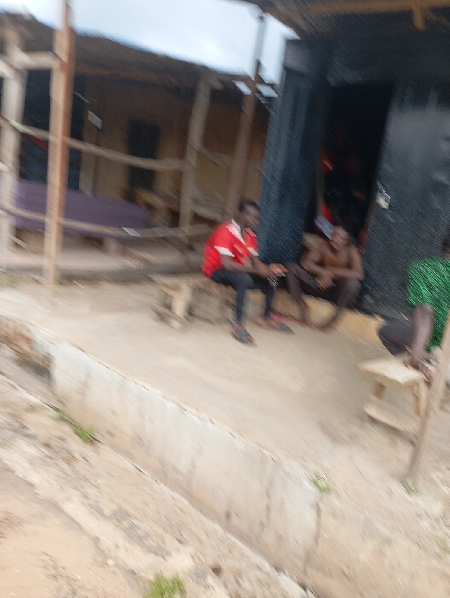 In Shendam, Nigeria, three men are captured in a moment of repose. The first man, on the left, is seated on a low, rough bench, wearing a red and white shirt and dark pants. He holds a dark object in his right hand, possibly a phone. To his right, a second man, shirtless and with dark skin, sits on the ground with his legs crossed. He appears to be looking down. A third man, partially visible on the far right, is seated on a small, light-colored wooden stool, wearing a green shirt with a patterned design. He is seen from the waist down. The scene unfolds in front of a rudimentary structure, characterized by wooden supports and dark corrugated metal roofing. The ground is a mix of dirt and concrete, with a noticeable drainage channel running through it. The overall impression is one of a casual gathering in an outdoor, utilitarian setting.