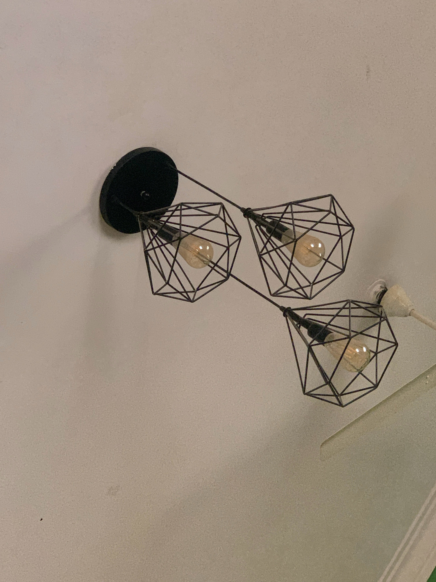 The image shows a modern black geometric chandelier with three Edison-style light bulbs hanging from a white ceiling. The chandelier is attached to a black circular base mounted on the ceiling. The light bulbs are illuminated, casting a warm glow. The ceiling appears to be textured and is painted a light color. A white cord is visible extending from one of the light fixtures. The photograph was taken from a low angle, looking up at the ceiling. The scene is indoors. The image's origin location is the city of Bengaluru, India.