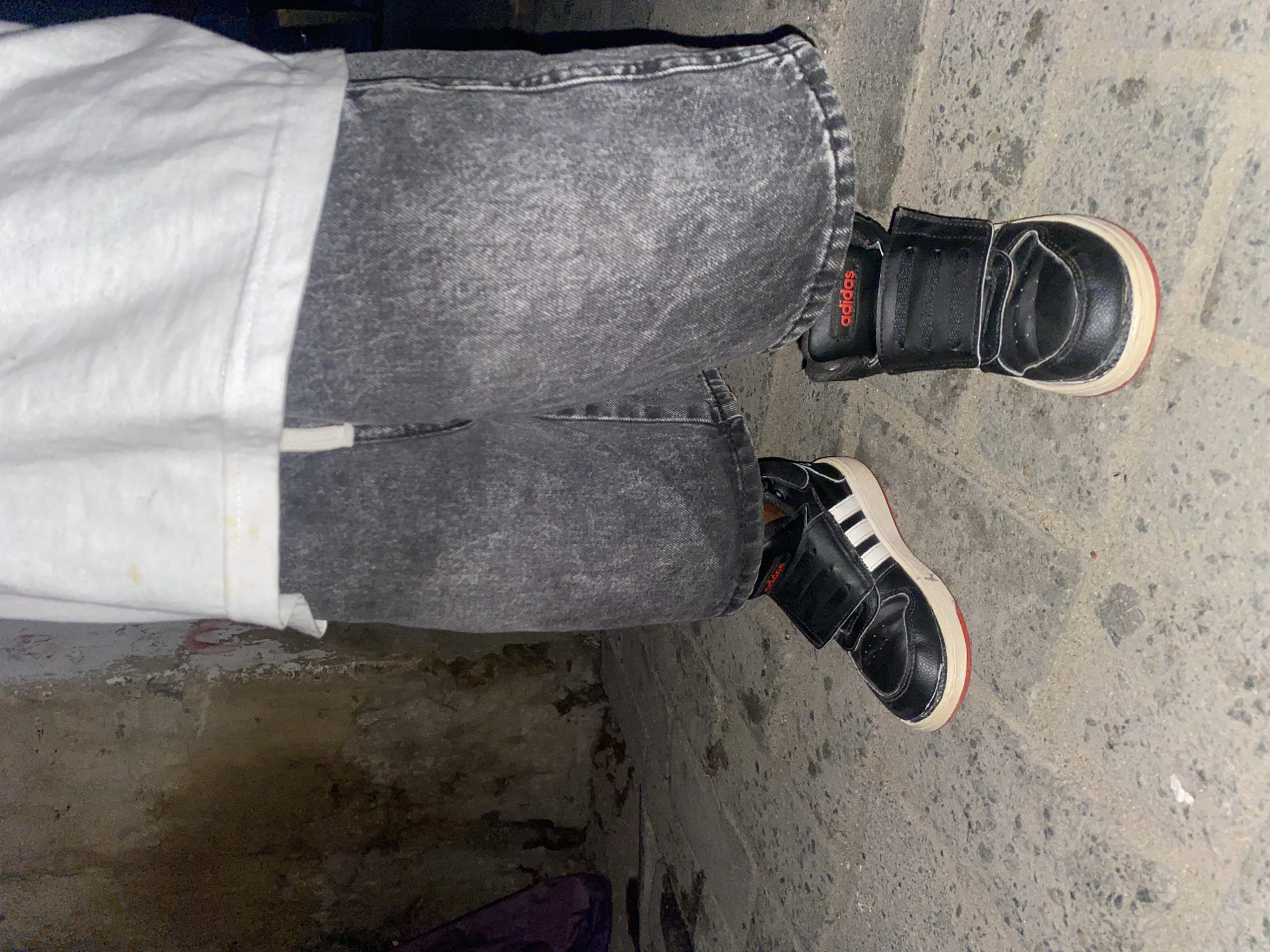 The image shows a person wearing grey acid-wash jeans, a white shirt, and two different black Adidas high-top sneakers. The left shoe has white stripes and a red sole, while the right shoe is plain black with a red Adidas logo. The person is standing on a concrete surface, likely outdoors. The location is possibly Ado, Nigeria. The ground appears somewhat soiled, suggesting a pedestrian area.