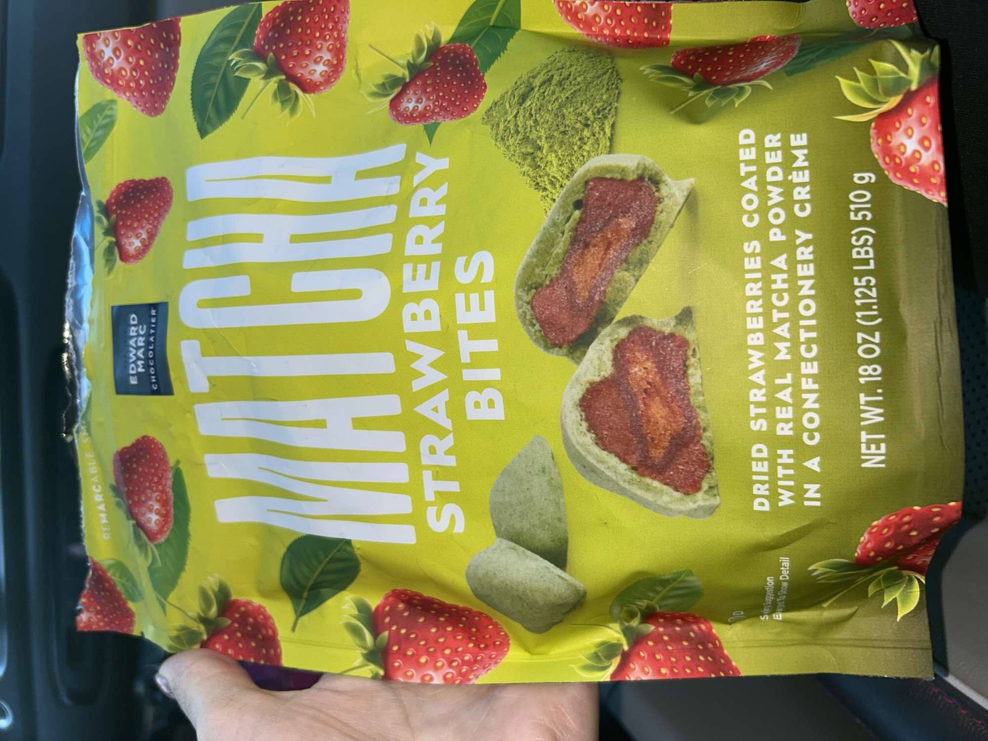 A person's hand is holding a bag of Edward Marc Chocolatier Matcha Strawberry Bites in Everett, Washington, USA. The bag is bright yellow and features images of strawberries, green leaves, and matcha powder. Text on the bag indicates "MATCHA STRAWBERRY BITES" and "DRIED STRAWBERRIES COATED WITH REAL MATCHA POWDER IN A CONFECTIONERY CREME". The weight is listed as "NET WT. 18 OZ (1.125 LBS) 510 g". The background appears to be the interior of a car.