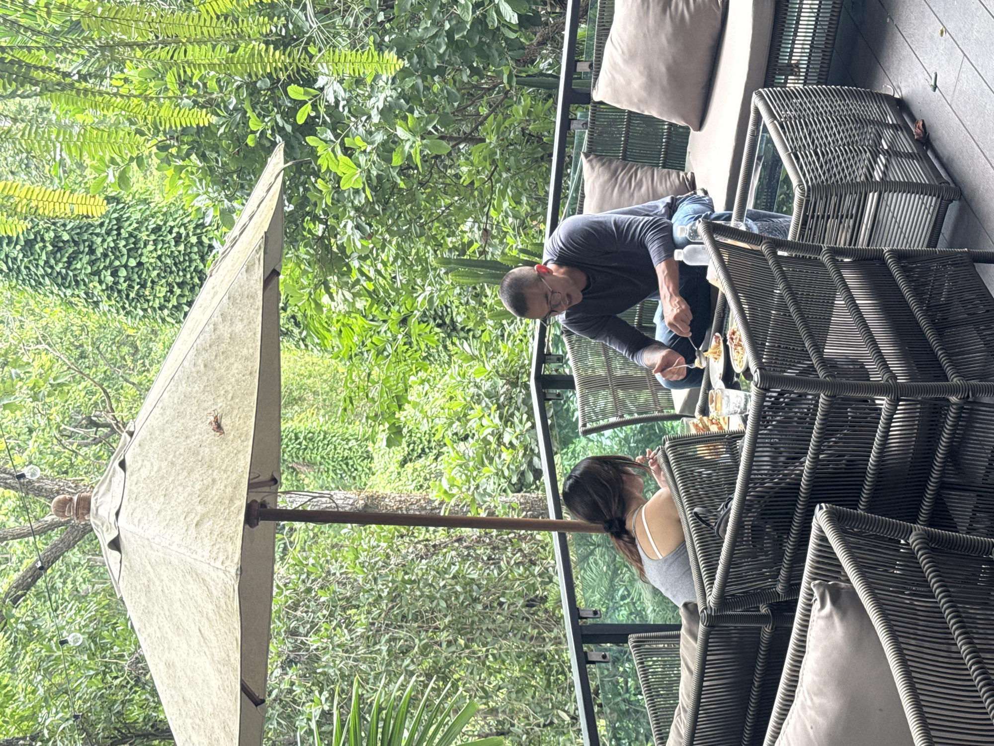 A man and a woman are seated at a table on an outdoor patio, beneath a large umbrella, amidst lush green foliage. The woman, on the left, is wearing a grey tank top and is looking down at her food, appearing to eat. The man, on the right, is wearing a grey long-sleeved shirt and blue jeans, and is actively eating, holding a fork with pasta. A glass of water and a bottle of water are visible on the table. The patio furniture is made of woven wicker, with plush cushions. The background is dominated by dense green trees and plants, suggesting a tropical setting. The location is identified as Suthep, Thailand.