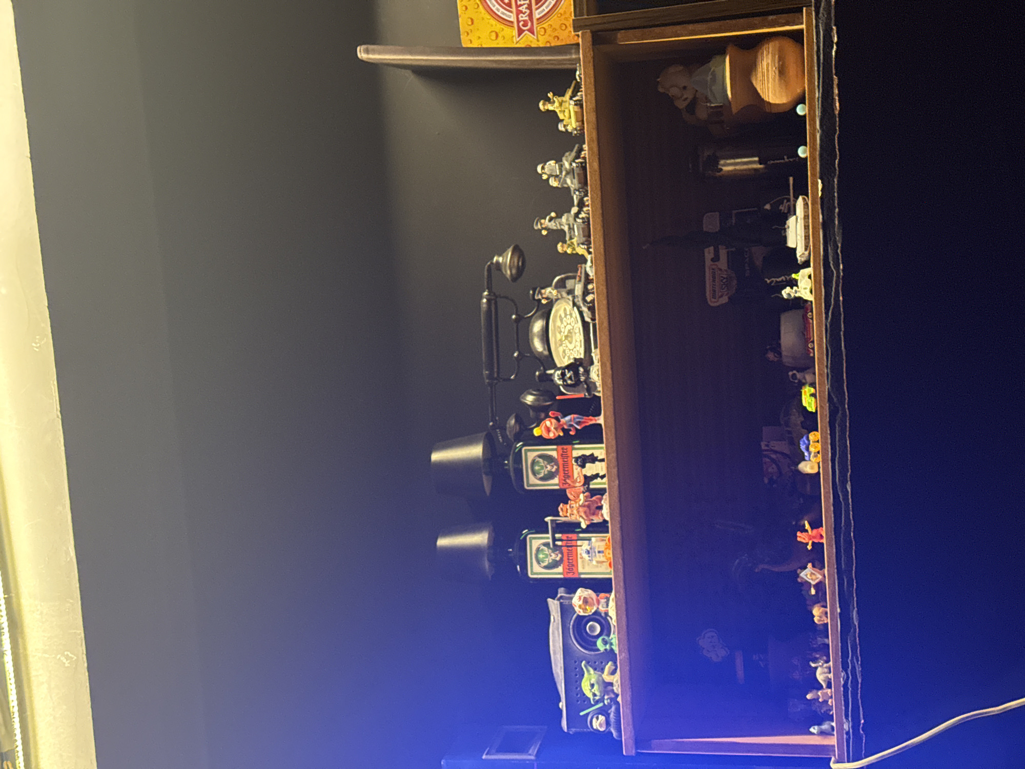The image shows a dark room with a wooden shelf mounted on a dark gray wall. The shelf is filled with various objects, including figurines, a toy telephone, and a bottle of Jägermeister. The lighting is dim, with a bright light source coming from the left side of the frame. The location information (Yerevan, Armenia) cannot be verified from the image alone.