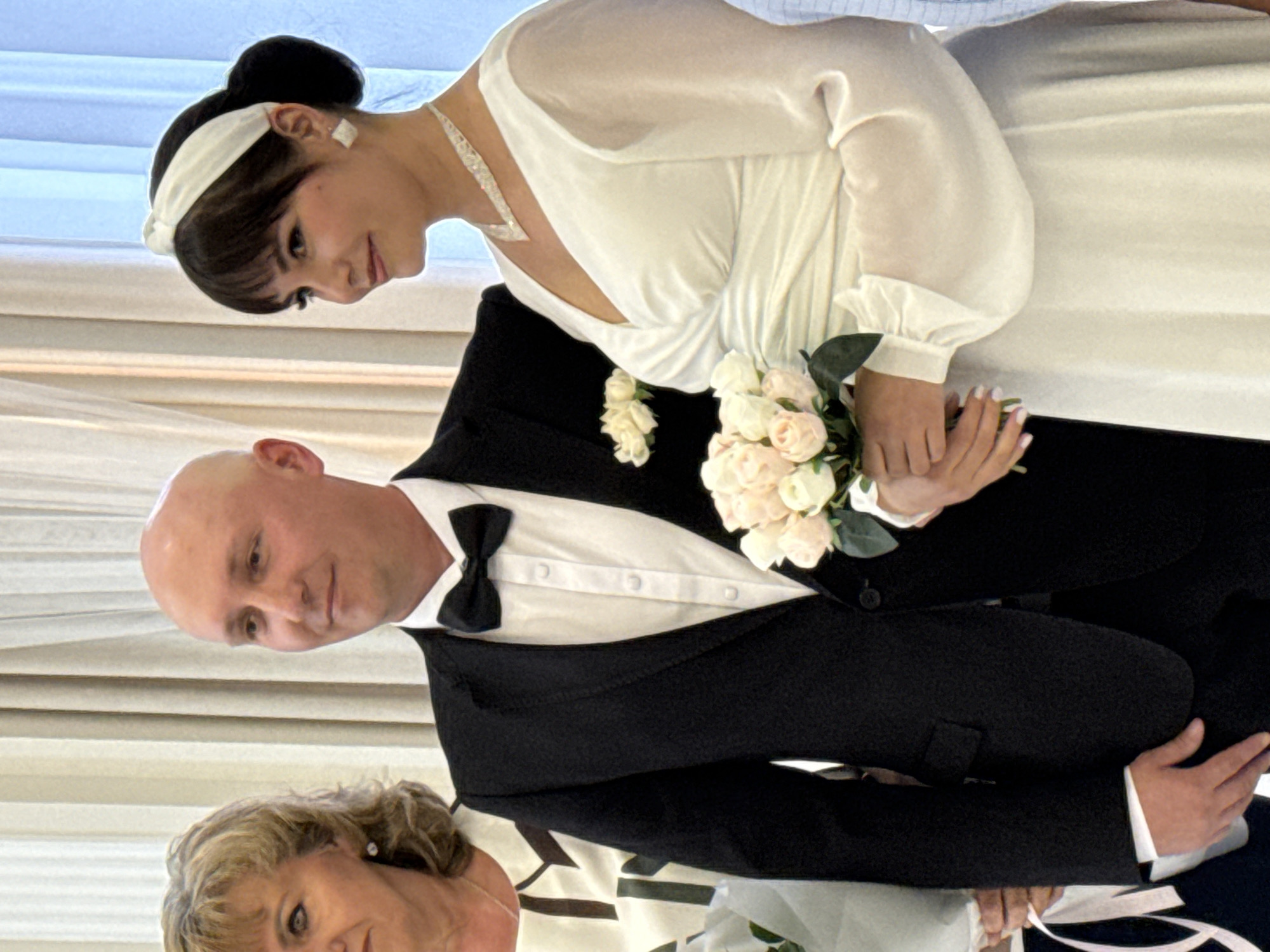 A wedding photo taken in an indoor location, possibly a venue or a home, in Unknown, Unknown. The image features a man and a woman, presumably the bride and groom, standing side by side. The man is wearing a black tuxedo with a white shirt and a black bow tie. He is bald and has a light complexion. The woman, dressed in a white wedding gown, has dark hair pulled back with a white headband and is wearing a diamond necklace and earrings. She is holding a bouquet of light-colored roses. To the left of the couple, a woman, likely a mother or a guest, is partially visible. She has blonde hair and is wearing a dress with a patterned neckline. The background consists of neutral-colored curtains.