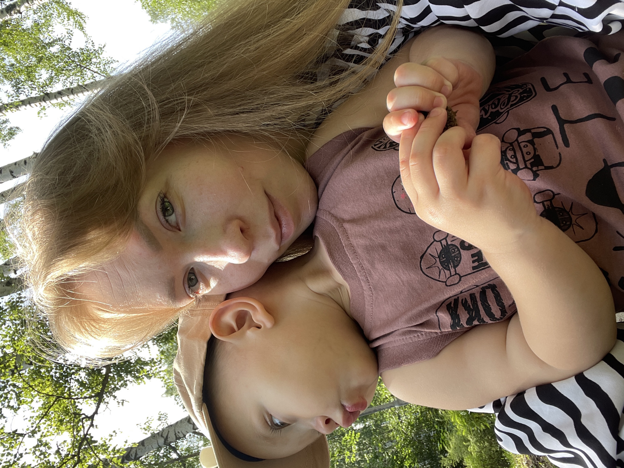 This is a close-up, selfie-style photograph featuring a woman and a young child outdoors, surrounded by trees. The woman, with blonde hair, is in the foreground, her face partially obscured as she looks towards the camera. The child, wearing a beige hat and a mauve t-shirt with graphics, is nestled against the woman, turned to the side with a pout. The child's hands are visible, one holding a small, dark object that appears to be a pinecone or burr. The background consists of lush green foliage and birch trees, suggesting a natural setting. There are no identifiable landmarks or clues that would indicate the location as the city of Perm, Russia.