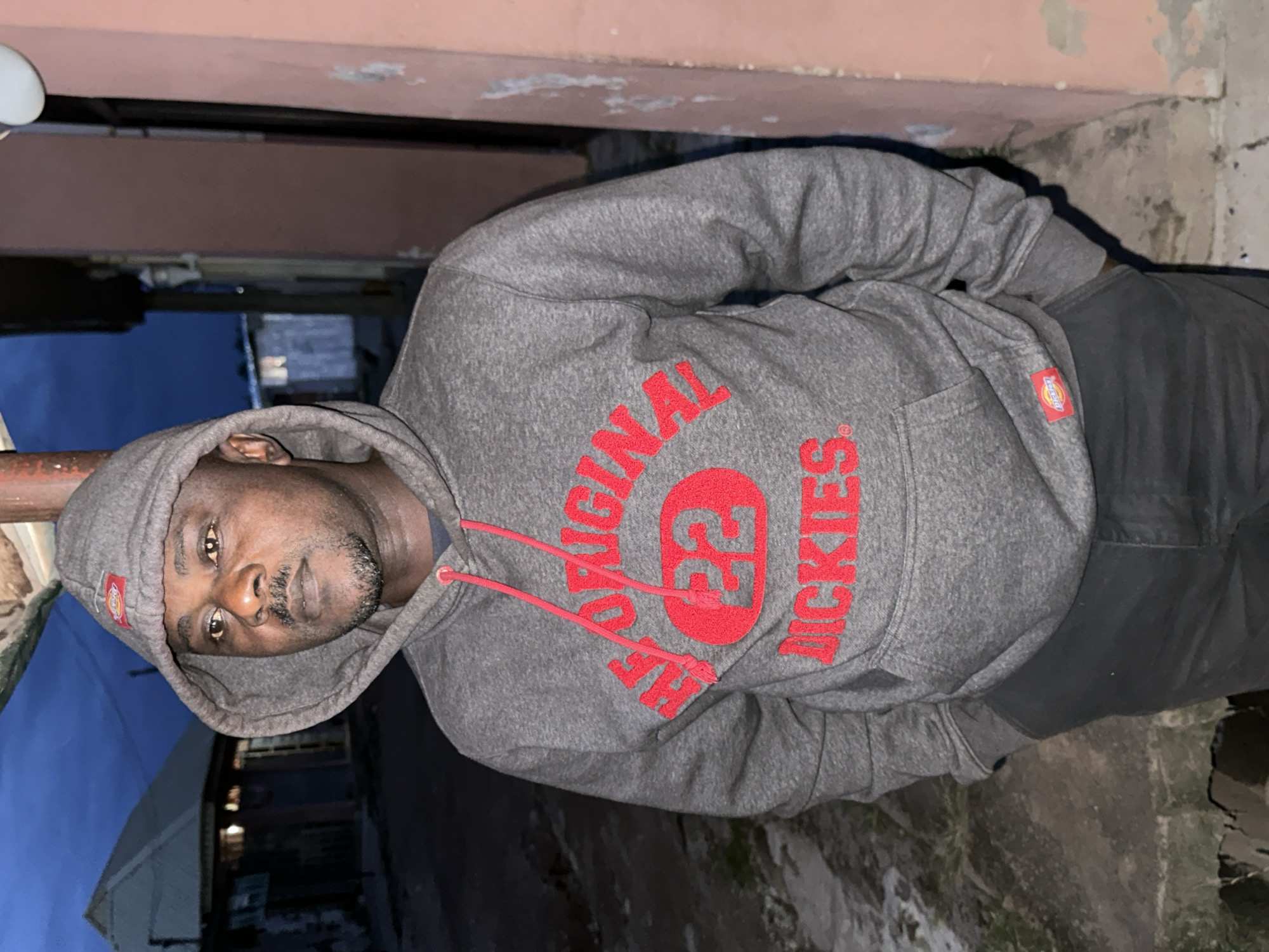 A man stands outdoors in Narayi, Nigeria, against a background of buildings and a twilight sky. He is wearing a grey hooded sweatshirt with red lettering that reads "THE ORIGINAL 22 DICKIES". His hands are in the front pocket of his sweatshirt. The man has a mustache and a goatee. The environment appears to be a residential or commercial area with some construction visible. The lighting suggests it is either early morning or late evening.