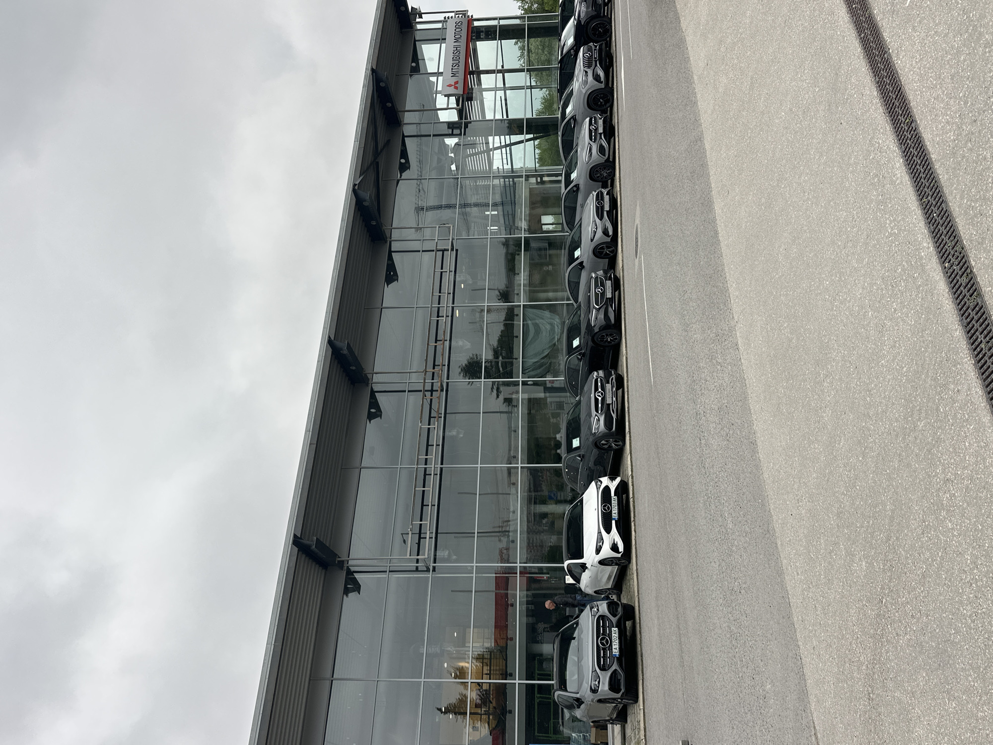 The image shows the exterior of a modern car dealership building under an overcast, cloudy sky. The building features a striking glass facade that reflects the surrounding environment, including trees and the pale sky. A prominent vertical sign displaying "MITSUBISHI MOTORS" is mounted on the upper part of the glass front.
Lined up along the pavement in front of the building are several cars, predominantly SUVs. These vehicles, in shades of white, grey, and dark grey/black, appear to be Mercedes-Benz models, identifiable by the star logo on their grilles, contrasting with the Mitsubishi branding on the building. Visible license plates on some of the cars, such as "EA 1701 AK," "EA 1702 AK," and "EA 1703 AK," indicate their registration in Varna, Bulgaria.
The foreground shows a paved asphalt surface with a long, linear drainage grate running parallel to the building and the parked cars. The scene depicts a daytime setting with diffused lighting due to the cloud cover. A faint reflection of a person can be seen in the glass facade near one of the white cars, suggesting potential activity inside or nearby. The cars appear to be part of the dealership's sales inventory.