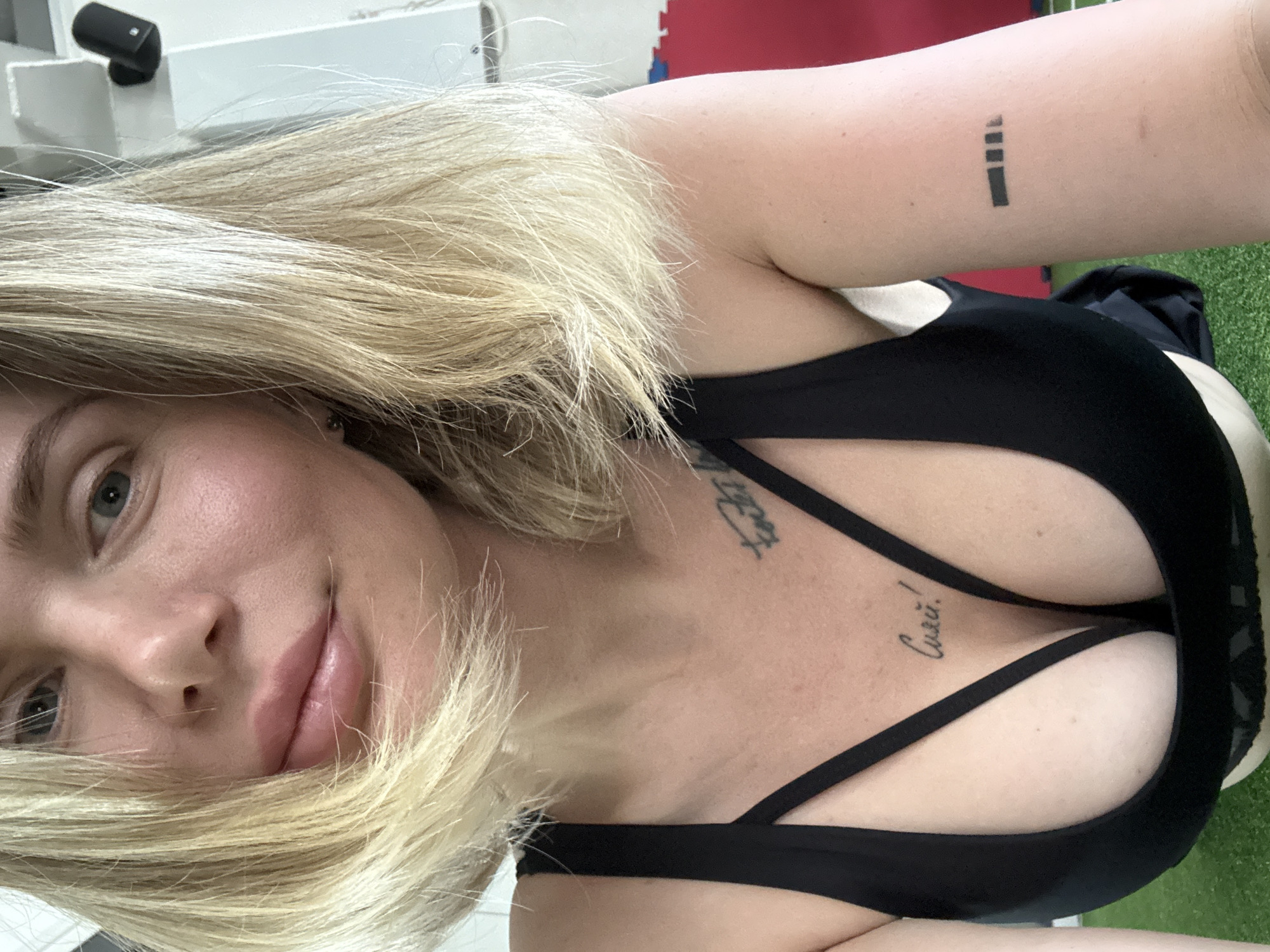 A close-up, selfie-style shot captures a woman with blonde hair, wearing a black sports bra, in what appears to be an indoor gym. Her gaze is directed towards the camera. The background shows a white wall with a black speaker mounted on it, and a portion of a red and blue mat on the floor. The woman has tattoos on her chest and right arm. There is no indication of the location being in Mayma, Russia.