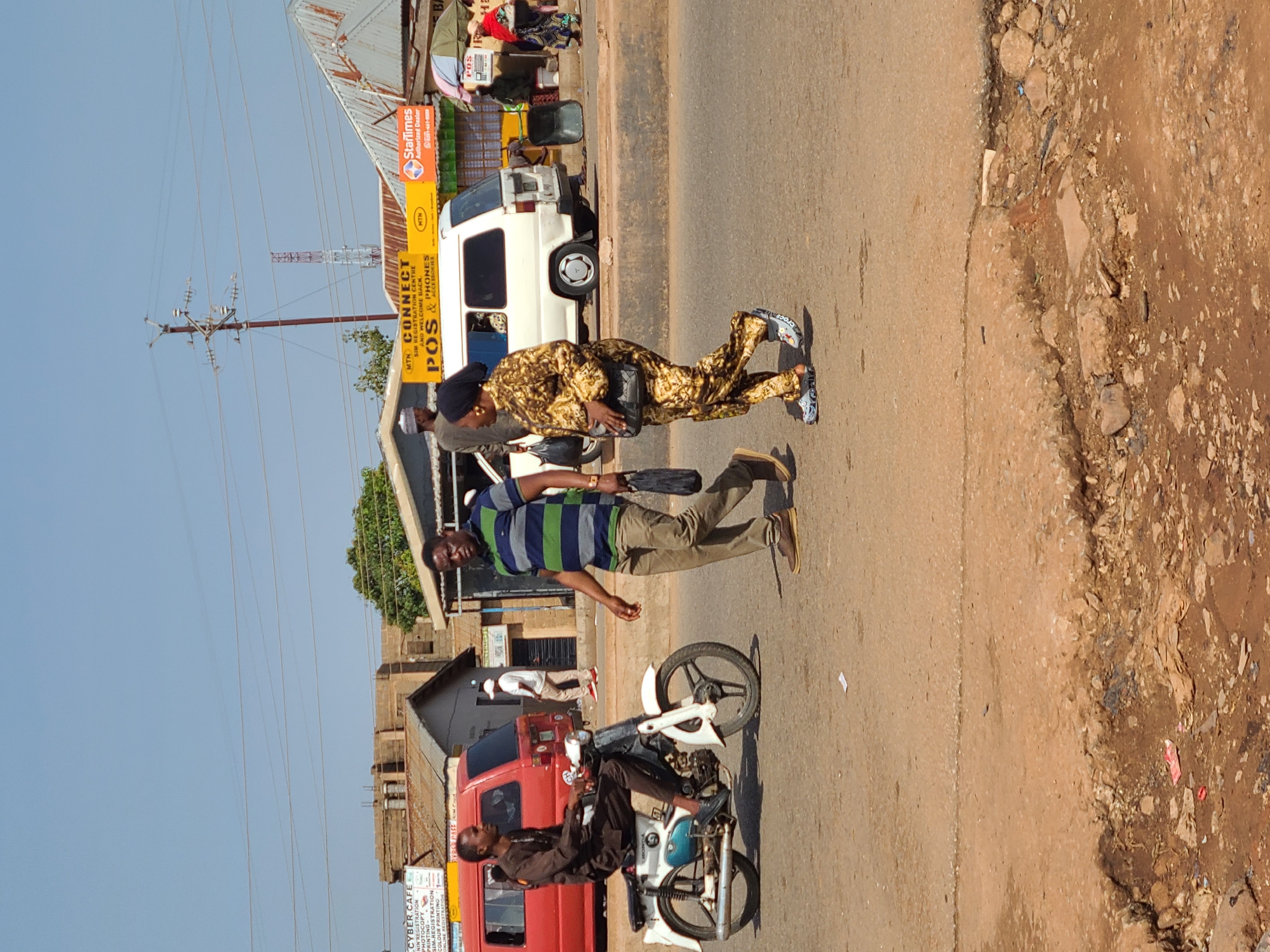 In Gura Top, Nigeria, a man in a brown traditional outfit rides a white Honda motorcycle in the foreground, facing left. To his right, a man in a striped shirt and khaki pants walks across the road, carrying a black bag. Further to the right, a woman in a patterned yellow and brown outfit also walks across the road, carrying a black purse and wearing blue Crocs. In the background, a white van is parked. To the left of the van, a red van is visible. Various shops and buildings line the street, including one with a prominent yellow sign reading "MTN CONNECT POS & PHONES" and another with an orange "StarTimes Authorized Dealer" sign. The road is made of asphalt and appears worn, with some areas of exposed dirt and rocks on the side. The sky is clear and blue.