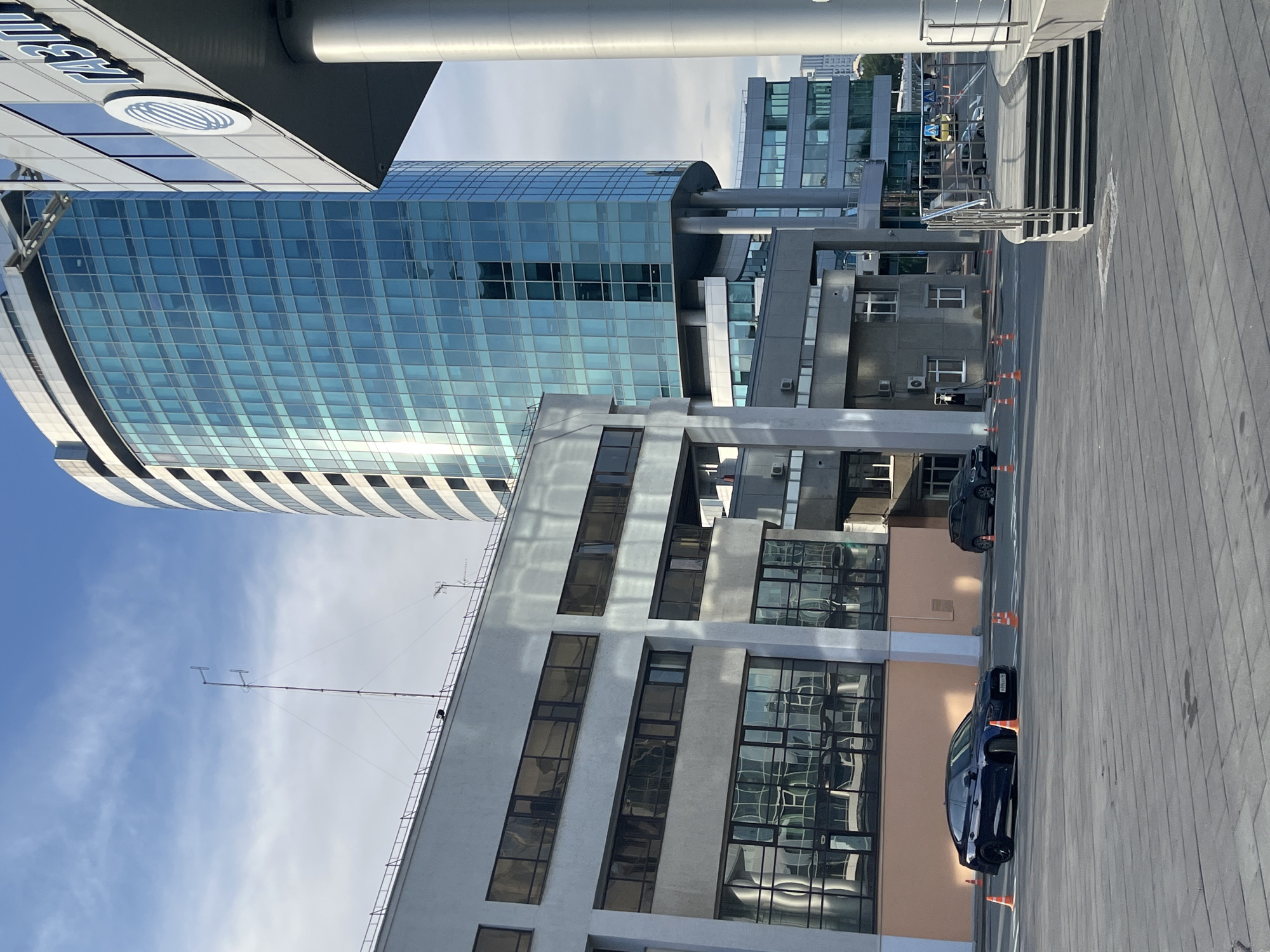 An exterior, eye-level, medium shot shows a wide, paved area in front of several modern office buildings under a partly cloudy sky. In the foreground, the gray paving stones are laid in a pattern. To the left of the frame, a dark blue sedan is parked in the paved area. Orange traffic cones are placed around the sedan. Further back, a darker colored SUV is also parked. In the midground, a low, rectangular building with large, dark-tinted windows runs horizontally across the frame. Behind it and to the right, a tall, cylindrical skyscraper with a blue glass facade rises prominently. A white and gray building with a large circular logo and the word "ГАЗПРОМ" in Cyrillic is partially visible to the right of the skyscraper. Additional, smaller office buildings with glass windows are visible in the background, some of which are lighter in color. The environment appears to be a commercial district.