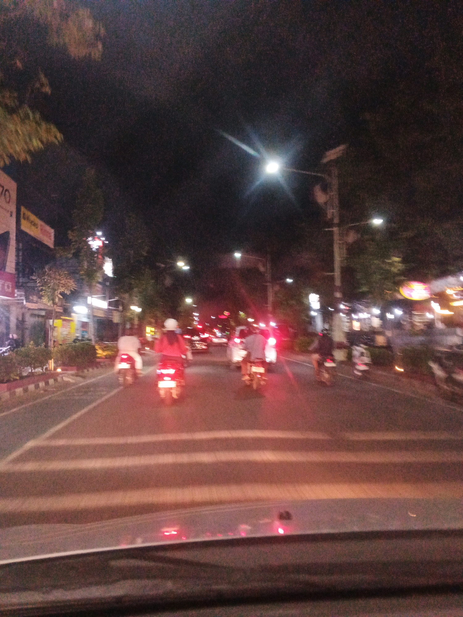 The image captures a night scene on a street in Jatisari, Indonesia. A road with clear lane markings is visible, with several motorcycles riding in the same direction. The motorcycles are illuminated by their headlights and taillights, with the taillights of the closest motorcycles casting a red glow on the road. Buildings and trees line the street, with streetlights providing illumination. Some signs are visible on the buildings, though their text is unreadable due to the low light and motion blur. The foreground shows the dashboard of a vehicle, indicating the photo was taken from inside a car. The overall impression is a typical urban night scene with moderate traffic.