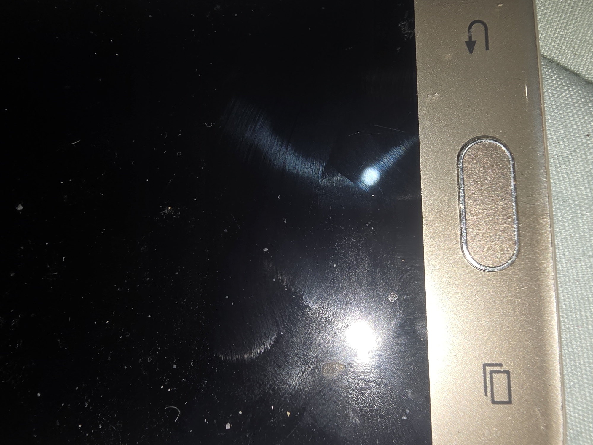 The image shows a close-up of a gold-colored smartphone with a black screen. The screen is dusty and has visible smudges and scratches, indicating it may not be new. There are bright light reflections on the screen, possibly from a light source. At the bottom of the phone, there are three buttons. The leftmost button has an icon resembling two overlapping rectangles, suggesting a recent apps or multitasking function. The middle button is a physical home button, oval-shaped and recessed, with a metallic finish. To the right of the home button is another icon, a curved arrow, which typically signifies a back function. The phone is resting on a light blue textured surface, possibly a cloth or fabric. The image is a still photograph and does not depict any activity or specific location within Ulan-Ude, Russia.