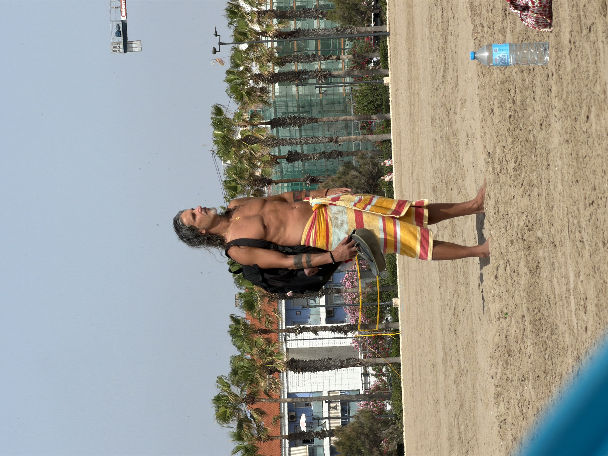 A man stands on a sandy beach, wearing a colorful striped towel wrapped around his waist and carrying a black backpack. He holds a dark-colored flip-flop in his right hand. His body is bare from the waist up, revealing tattoos on his left arm. He has graying hair and a beard. Behind him, palm trees line a beach with a volleyball net. Further in the background, buildings with orange roofs and blue facades are visible, with some structures undergoing construction, covered in green netting. The sky is clear and blue. A water bottle and a patterned bag lie on the sand to the right of the man.