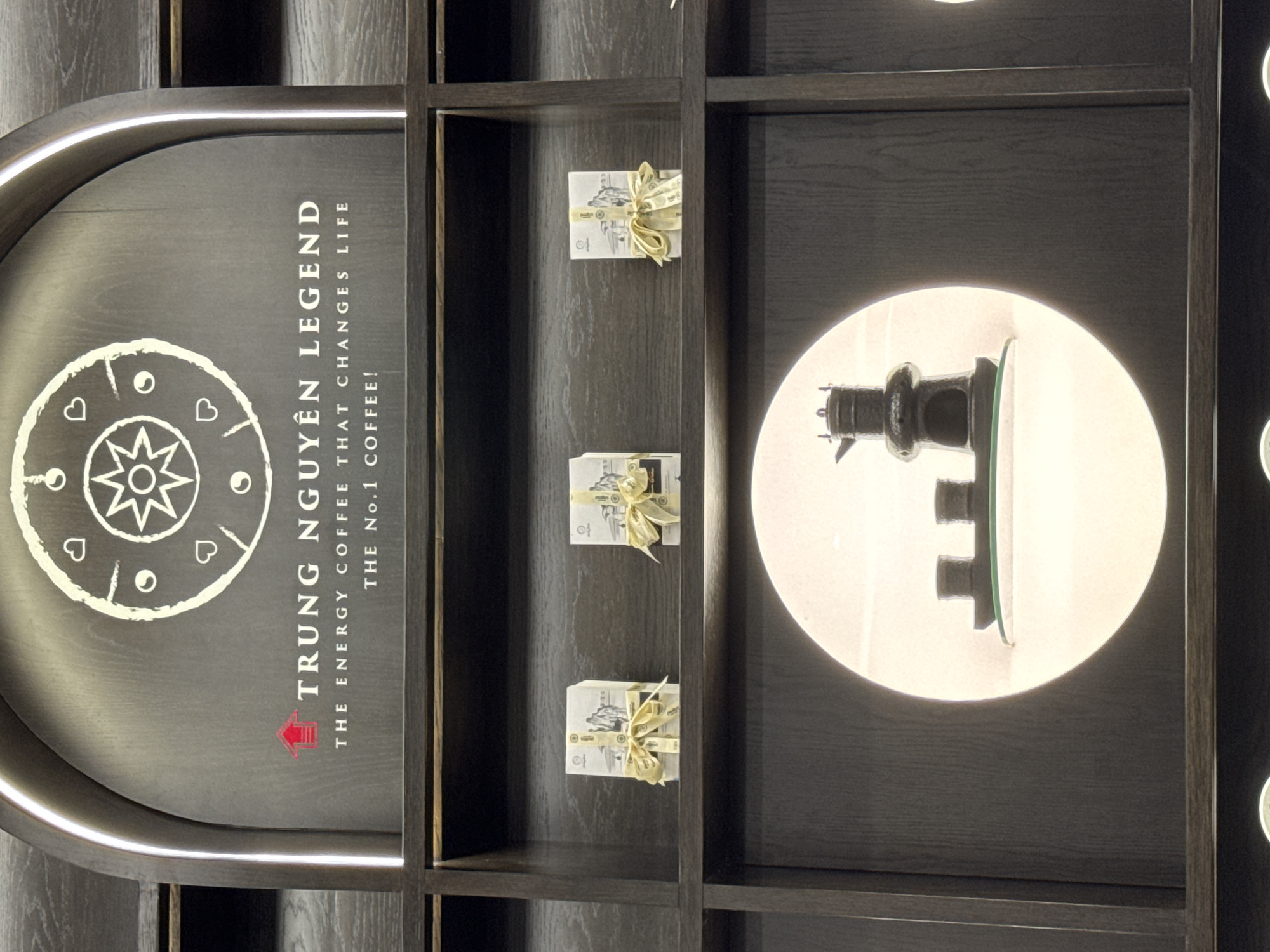 The image displays a section of an interior, likely a retail space, featuring a dark wooden shelving unit with various products. At the top, a large, dark gray arched panel displays white text that reads "TRUNG NGUYEN LEGEND" followed by "THE ENERGY COFFEE THAT CHANGES LIFE THE No.1 COFFEE!". Above the text is a white circular emblem with a star-like design in the center, surrounded by smaller circular motifs and heart shapes.
On the shelf directly below the panel, three small, rectangular packages are displayed, each wrapped in white paper and tied with a yellow ribbon. These packages appear to be gift-wrapped coffee products.
Further down, a circular illuminated display niche contains a dark, ornate coffee pot and two small cups, all resting on a clear glass shelf. The niche has a soft, diffused lighting.
The shelving unit itself is constructed of dark wood with visible grain. The overall aesthetic suggests a modern and sophisticated presentation of coffee-related items. The scene is indoors, with no visible windows or outdoor elements. The location is identified as Trung Nguyen Legend Cafe, 76 Tran Phu Street, Nha Trang Ward, Vietnam, which is a prominent coffeehouse chain in Vietnam.