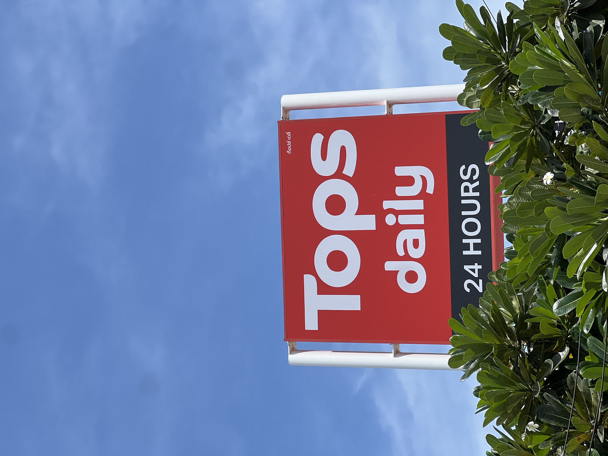 A low-angle shot of a "Tops daily 24 HOURS" sign in Unknown, Unknown. The sign is red and black, with white lettering. It is mounted on a white metal frame. The sign is partially obscured by green leafy tree branches. The sky is blue with some wispy clouds.