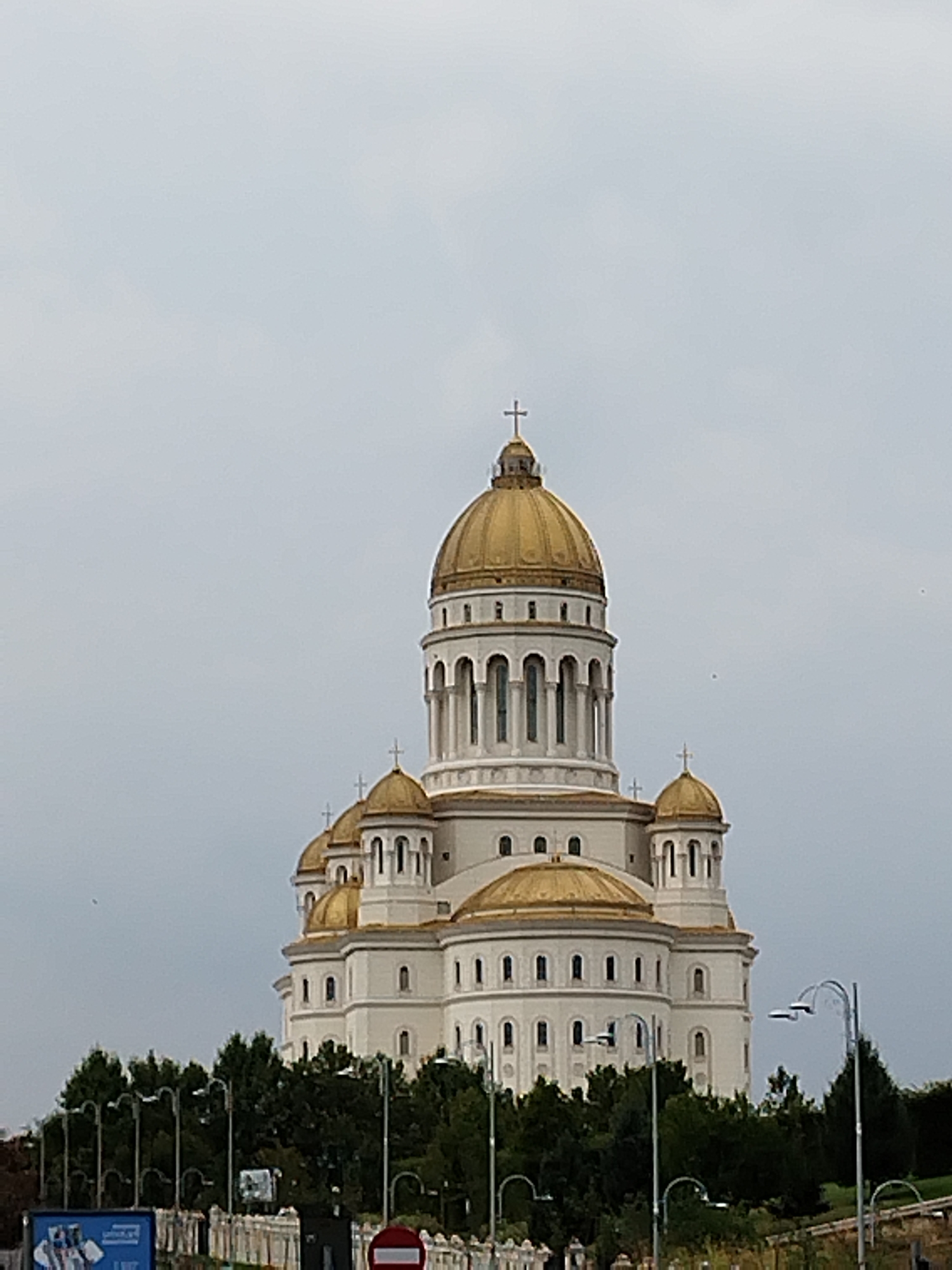 The photograph depicts a large, ornate cathedral with multiple golden domes, characteristic of Orthodox architecture, set against a cloudy, overcast sky. The cathedral is white with classical columns and arched windows, featuring crosses atop its domes. In the foreground, a line of dark green trees obscures the lower portion of the structure. Several streetlights with modern designs are visible in front of the trees. A fence runs horizontally across the bottom of the image, with glimpses of what appear to be street signs and a billboard. This is the Patriarchal Cathedral of Romania, located in Bucharest, Romania.