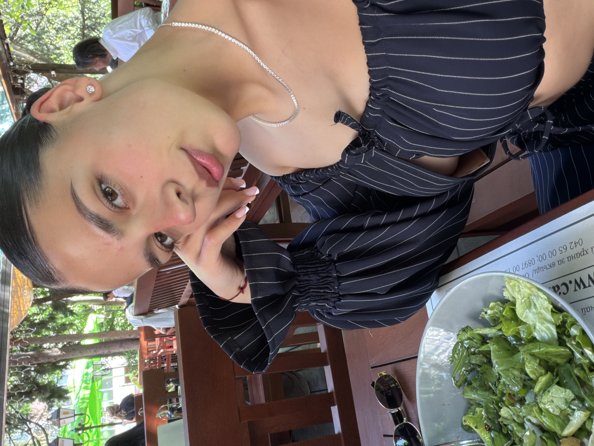 A woman with olive skin and light-colored eyes wearing a thin silver chain and a dark blue and white pinstriped top sits at a wooden table outdoors. Her left hand is resting on her face. To her right is a plate of green salad and a pair of sunglasses. The setting appears to be a restaurant in Stara Zagora, Bulgaria, as indicated by the text on the paper near the plate: "www.ca... хранa за вкъщи / O... 042 65 00 00; 0897 00 15". Other people are visible seated at tables in the background among trees.