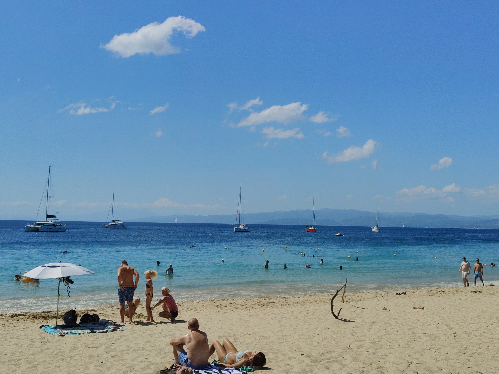 The image depicts a sunny day at a beach in an unknown location, with several people enjoying the water and sand. The sea is a vibrant blue, with a clear horizon meeting a bright blue sky dotted with white clouds. Various sailboats are visible in the distance, some closer to shore and others further out.
On the sandy beach, there are several groups of people. In the foreground, a man and a woman are lying on beach towels, relaxing. Further back, a family is gathered near a beach umbrella. A man in blue shorts stands with his back to the camera, while a young girl in a swimsuit stands beside him, and a younger child plays in the sand at their feet. Another woman in a pink top and dark shorts is crouching nearby.
Several other individuals are in the shallow water near the shore, some swimming and others wading. Yellow buoys are scattered in the water, marking a swimming area. The beach itself is sandy, with some scattered driftwood and a few pieces of luggage near the umbrella. The overall atmosphere is one of leisure and summer vacation.