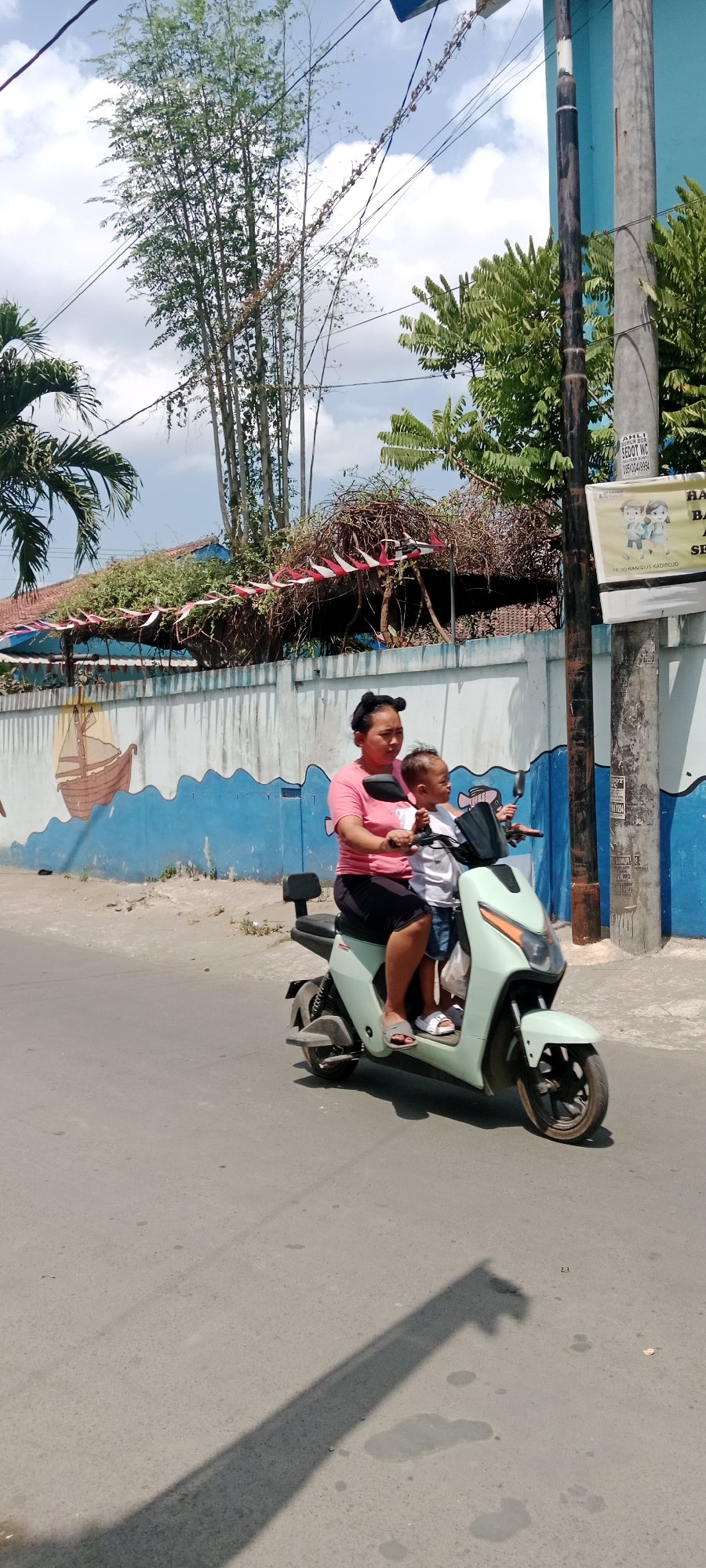 In Purwomartani, Indonesia, a woman rides a mint green electric scooter on a paved road. She is wearing a pink t-shirt and black skirt, with her hair tied up in two buns. A young child, wearing a white t-shirt and blue shorts, sits in front of her. The scooter has a black seat and a black windshield. Behind the scooter is a blue and white wall with a mural of a sailboat. The wall is decorated with red and white pennant flags. Lush green trees and vegetation line the background, with a clear blue sky above. Utility poles and wires are visible to the right of the frame.