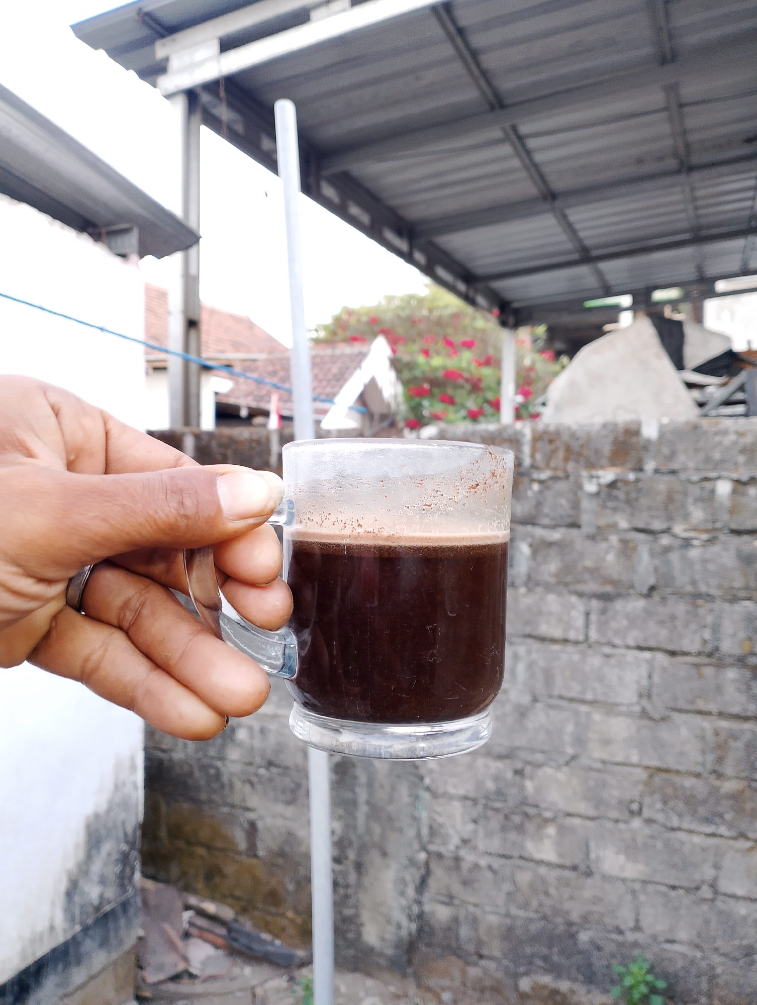 A hand holds a clear glass mug of dark liquid, likely coffee, in front of a rough, grey brick wall. The mug has a light brown foam on top. The hand has a silver ring on the ring finger. In the background, a corrugated metal roof is visible, supported by metal beams. Beyond that, the tops of houses with red tile roofs and some pink flowers can be seen. The scene appears to be outdoors, possibly in a backyard or an open area within a residential setting. The lighting suggests it is daytime.