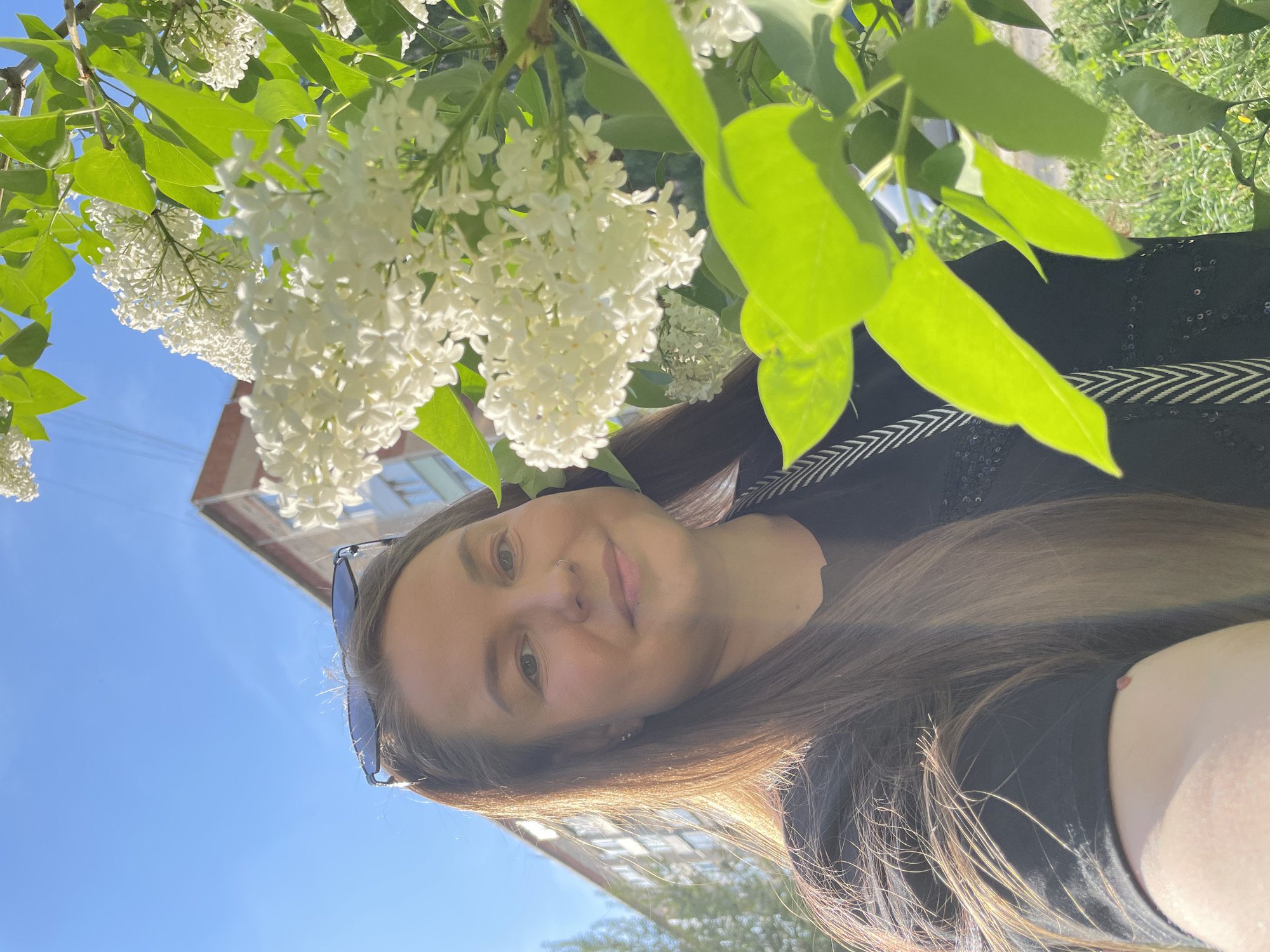 A woman is taking a selfie outdoors with white flowers in the foreground and a building partially visible in the background. The woman has long brown hair and is wearing a black shirt. She is smiling slightly. The flowers are white and appear to be lilacs, with green leaves visible among the blossoms. The sky is blue and clear. The location is Izhevsk, Russia, based on metadata, but this isn't clearly discernible in the image itself.