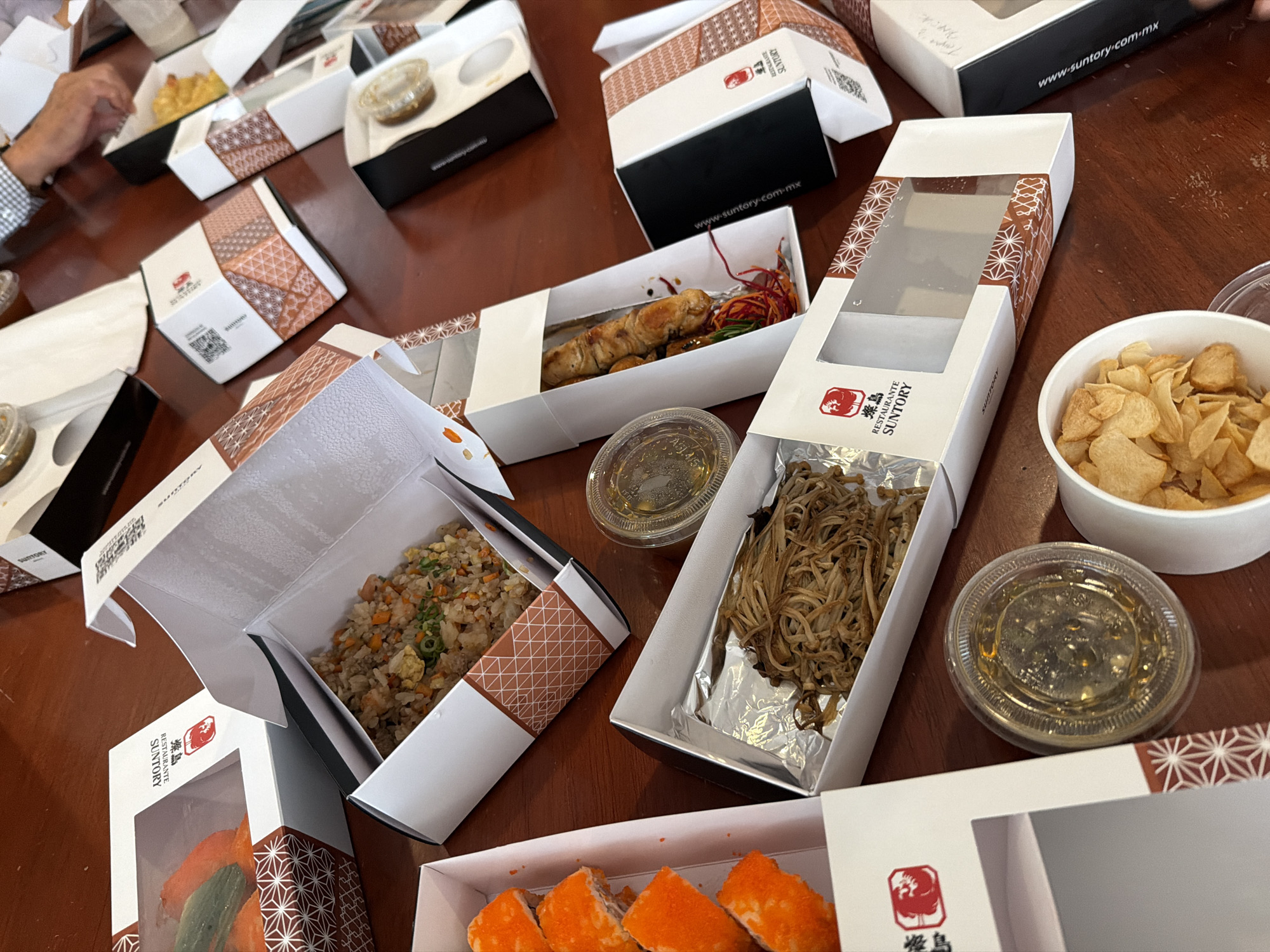 The image shows a table covered with various takeout containers from the Japanese restaurant Suntory. The containers are white with geometric patterned accents. Some of the containers are open, revealing different food items inside, including fried rice, yakitori skewers, noodles, sushi, and chips. There are also small clear containers containing sauces. The website "www.suntory-com-mx" is visible on some containers, indicating they are likely from a Suntory restaurant location in Mexico. The context and language suggest this scene is located in Mexico, possibly in the city of San Bernardino Tlaxcalancingo, Mexico, but there is no information in the image to confirm that.