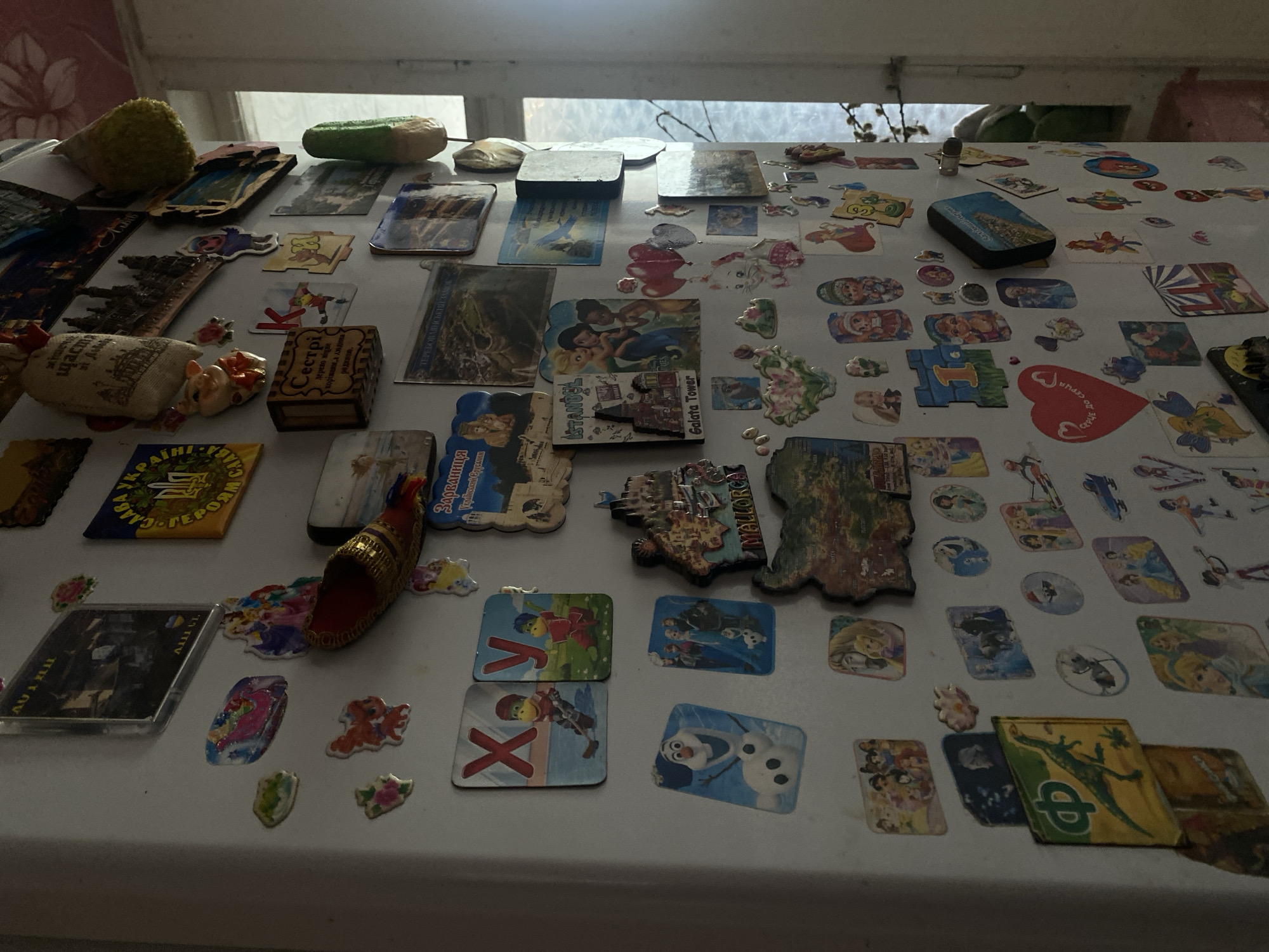 The image shows a cluttered refrigerator door covered with numerous magnets, stickers, and small decorative items. The magnets feature diverse themes, including landmarks from different cities (Istanbul, Galata Tower, "MELLORCA"), Ukrainian symbols and flags, cartoon characters (from "Frozen," "Ben and Holly's Little Kingdom"), and inspirational messages in Ukrainian. There's also a small wooden box with Ukrainian inscriptions. Other objects include decorative figures, a fake cucumber, and a souvenir shoe. The overall environment appears to be a domestic kitchen. The precise location in Pakhynia, Ukraine, cannot be determined.