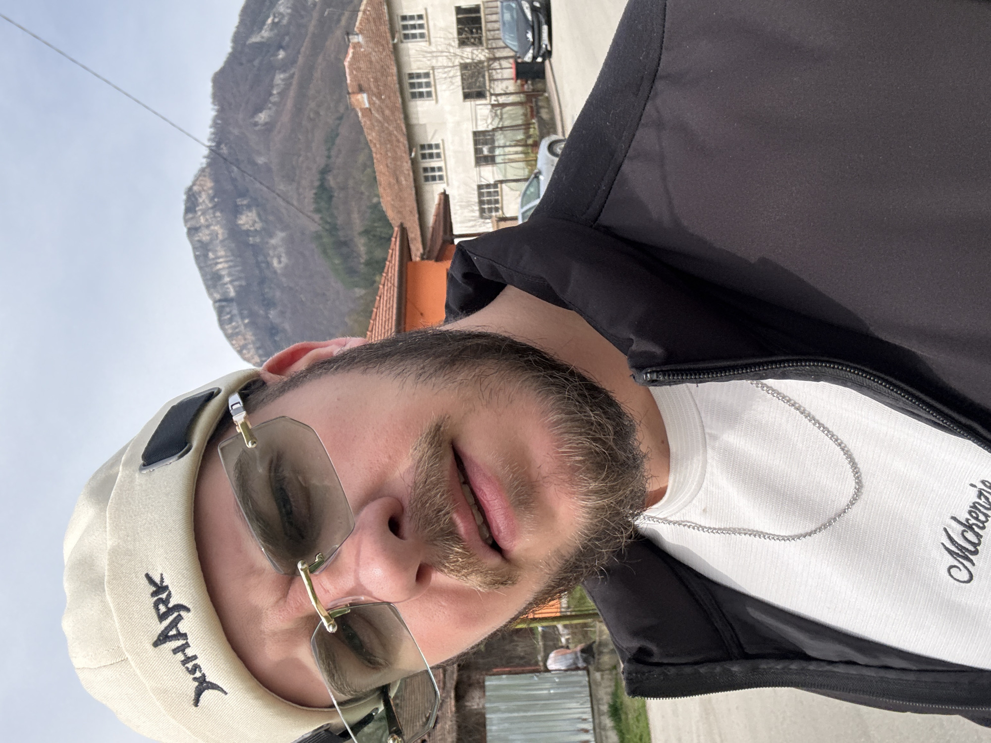 The image shows a selfie of a man wearing a cream-colored baseball cap with the word "Xshark" on it and aviator sunglasses. He is wearing a white shirt with the word "Mckenzie" written on it and a black jacket. A silver chain is visible around his neck.
Behind him, a building with multiple windows and a terracotta tiled roof can be seen. A mountain rises in the background. Cars are parked on the road. The scene appears to be set in the city of Teteven, Bulgaria.