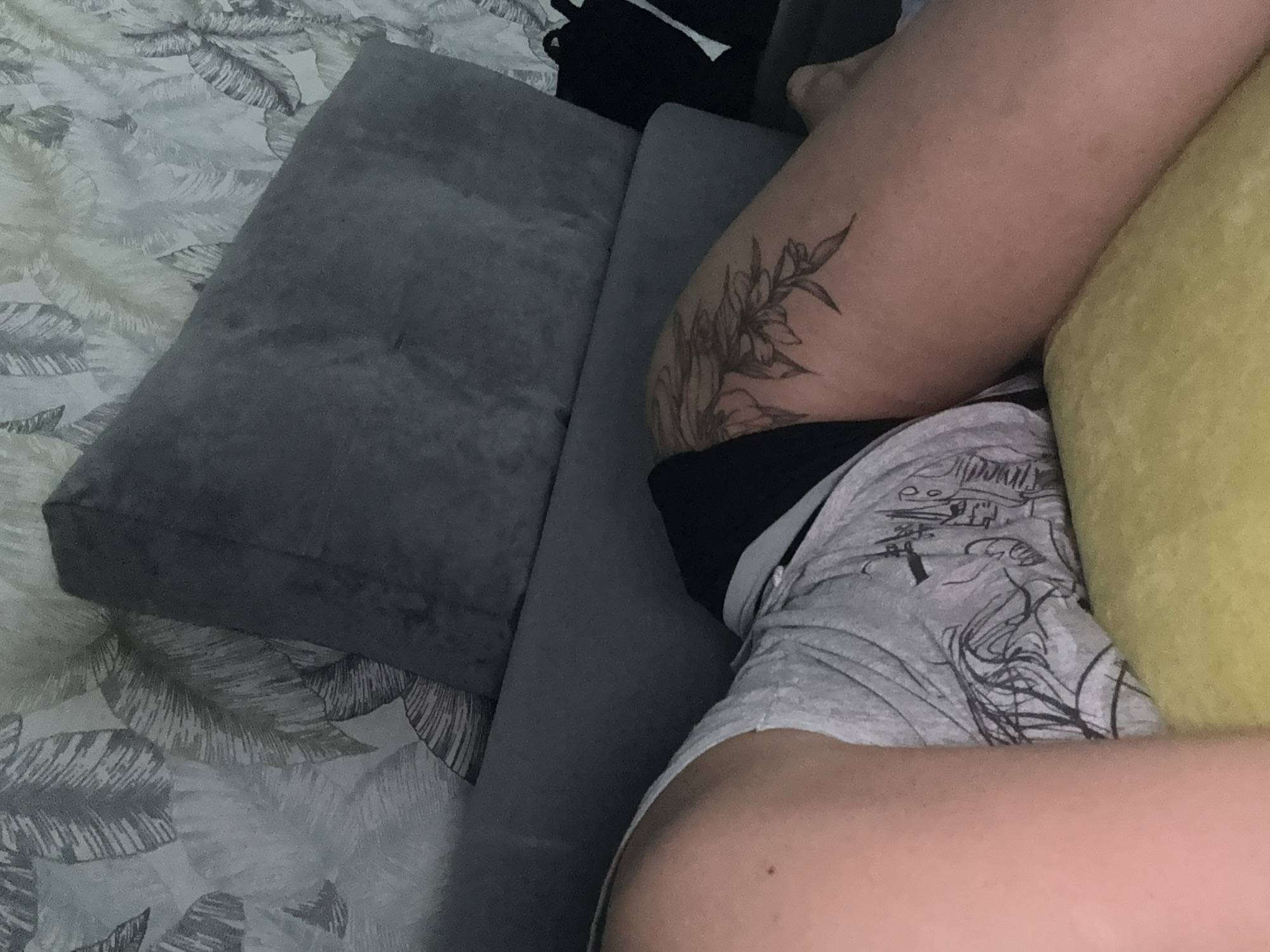 The image captures a close-up view of a person's legs and torso as they recline on a sofa or bed in an indoor setting. The scene is dominated by muted tones, with grey and white patterns, a grey sofa, and a yellow pillow providing some contrast.
A significant portion of the image shows the person's upper thighs and lower torso. One thigh, on the left side of the frame, features a prominent black line tattoo depicting floral or foliage elements. The person is wearing grey shorts with a black waistband, and the shorts have a dark, abstract graphic print that appears to include scribbled lines and possibly some unreadable text. A yellow pillow rests against the person's leg and hip on the right side of the frame.
The background features a grey upholstered sofa or couch, where the person appears to be resting. Beneath the sofa, and extending across the upper left and lower right of the image, is a bedspread or sheet with a distinctive grey and white pattern of large, palm-like leaves. The lighting in the room is subdued, suggesting an evening or dimly lit indoor environment. The location context provided is Kauchuk Stantsiya, Russia.