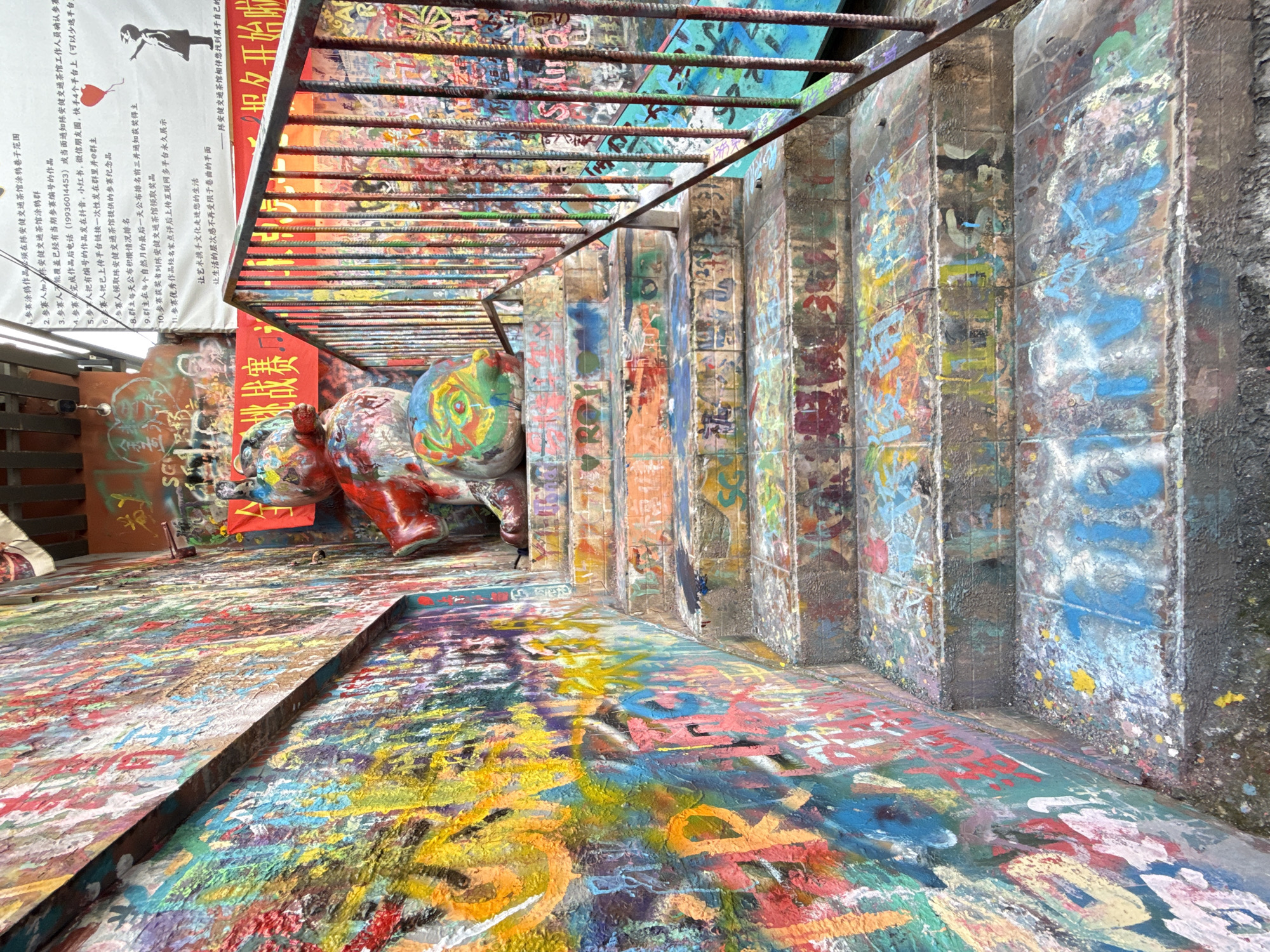 This image shows a colorful and artistic stairway in what appears to be the vicinity of the Chen Anjian Traffic Tea House in Jiulongpo District, China. The stairs and surrounding surfaces are covered with graffiti and paint in a vibrant array of colors. At the top of the stairs, there are sculptures of rhinos, also painted with colorful designs. Overhead, a metal grid or railing runs along the staircase. A sign with Chinese characters and an image of a figure holding a heart is visible, likely providing information or rules related to the artwork. The scene suggests a public art installation or project in the area.