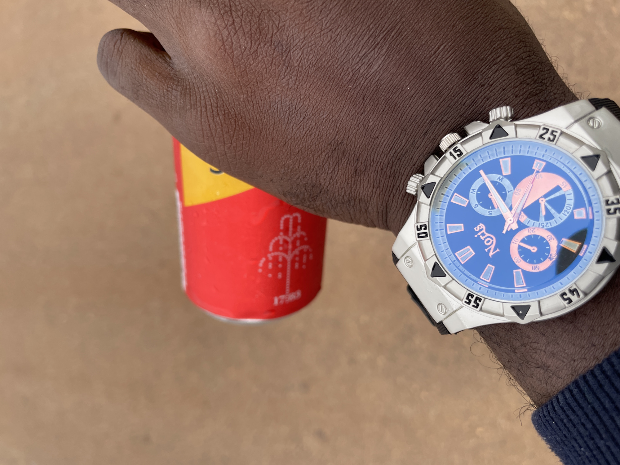 A close-up, eye-level shot shows a person's right arm and wrist wearing a silver and blue watch. The watch has a blue face with silver hands and markings, and a silver bezel with black triangular accents. The watch band is black. The person's arm is dark-skinned and hairy. In the background, slightly out of focus, is a red and yellow can, possibly a beverage can. The ground appears to be sandy or dusty. The lighting suggests it is daytime. The image was captured in Durumi, Nigeria.