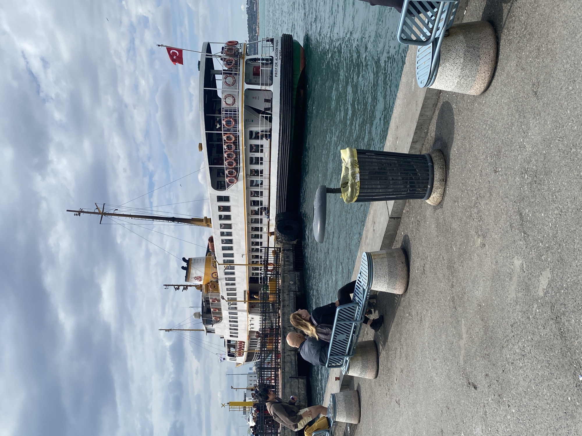 The image depicts a ferry docked at a pier in Kadıköy, Turkey. The ferry is white with a yellow stripe and a Turkish flag waving from its top. The name "PROF. DR. ALAEDDIN KIYICA ISTANBUL" is written on the side of the boat. The sky is overcast with gray clouds. On the pier, there is a black trash can next to the ferry dock. There are people sitting on benches. The pier is made of gray concrete.