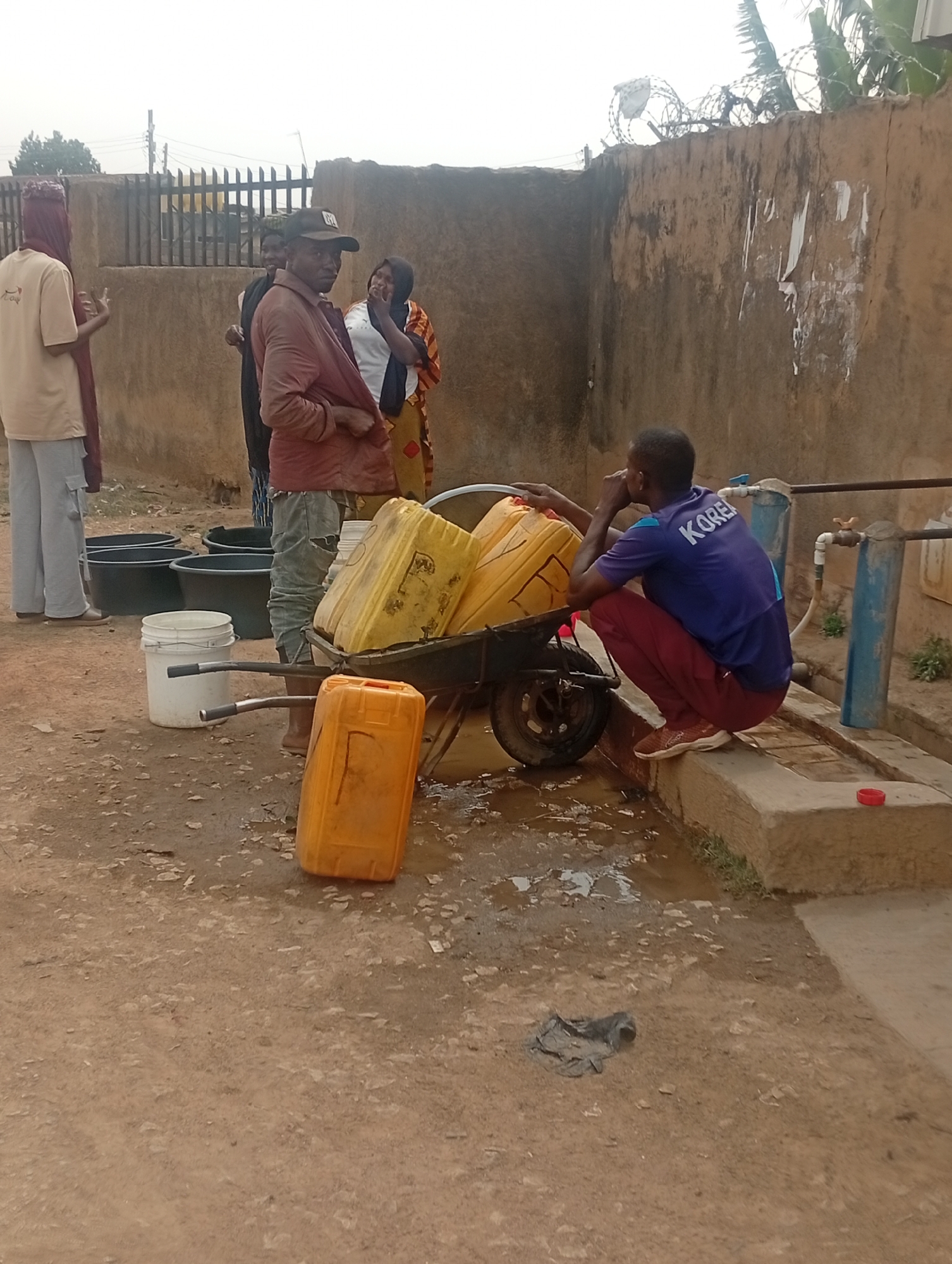 The image captures a bustling scene of water collection at a community point in Jos, Nigeria, under an overcast sky.
In the foreground, a man wearing a blue t-shirt with "KOREA" printed on the back and maroon pants squats beside a concrete platform, attending to a water tap and a blue water filtration system. A red cap rests on the concrete. Adjacent to him, a dark green wheelbarrow is heavily loaded with multiple yellow jerrycans, with one additional yellow jerrycan standing upright on the muddy ground. The area around the tap is visibly wet and muddy, indicating water spillage.
To the left, several large black plastic tubs and a white plastic bucket are placed on the ground, likely awaiting use for water collection. A group of people stands further back. On the far left, a person in a light-colored top, light pants, and a dark head covering is seen from behind, gesturing. Next to them, a man wearing a reddish-brown jacket and a baseball cap looks towards the viewer. Behind him, a woman in a patterned dress and white top, along with another individual, are engaged in conversation.
The background features a tall, rough-textured wall topped with coiled barbed wire, suggesting a secured boundary. Beyond the wall, a metal fence and some sparse foliage are visible under the hazy sky. The overall scene depicts a common activity of sourcing and transporting water in the region. Some faint markings, possibly the letter "F", are visible on the yellow jerrycans.
