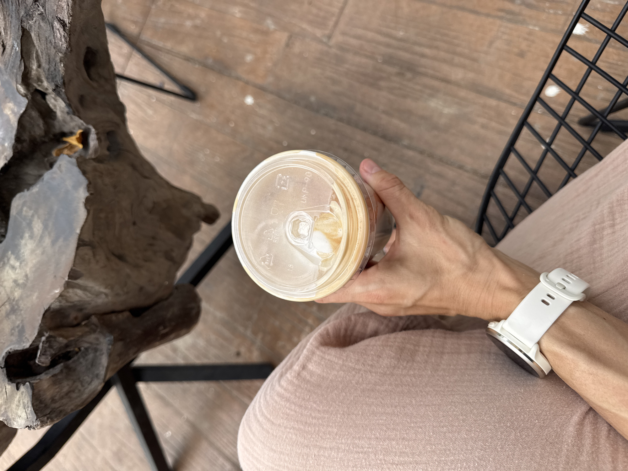 A top-down shot shows a person sitting outdoors, holding a plastic cup containing a beverage. The person wears a white smartwatch. The background consists of a wooden table, a metal chair, and a light pink garment, likely clothing. The image suggests a casual setting, potentially a cafe or outdoor seating area. There is no information available that confirms this photo was taken in Bac Nha Trang Ward, Vietnam.