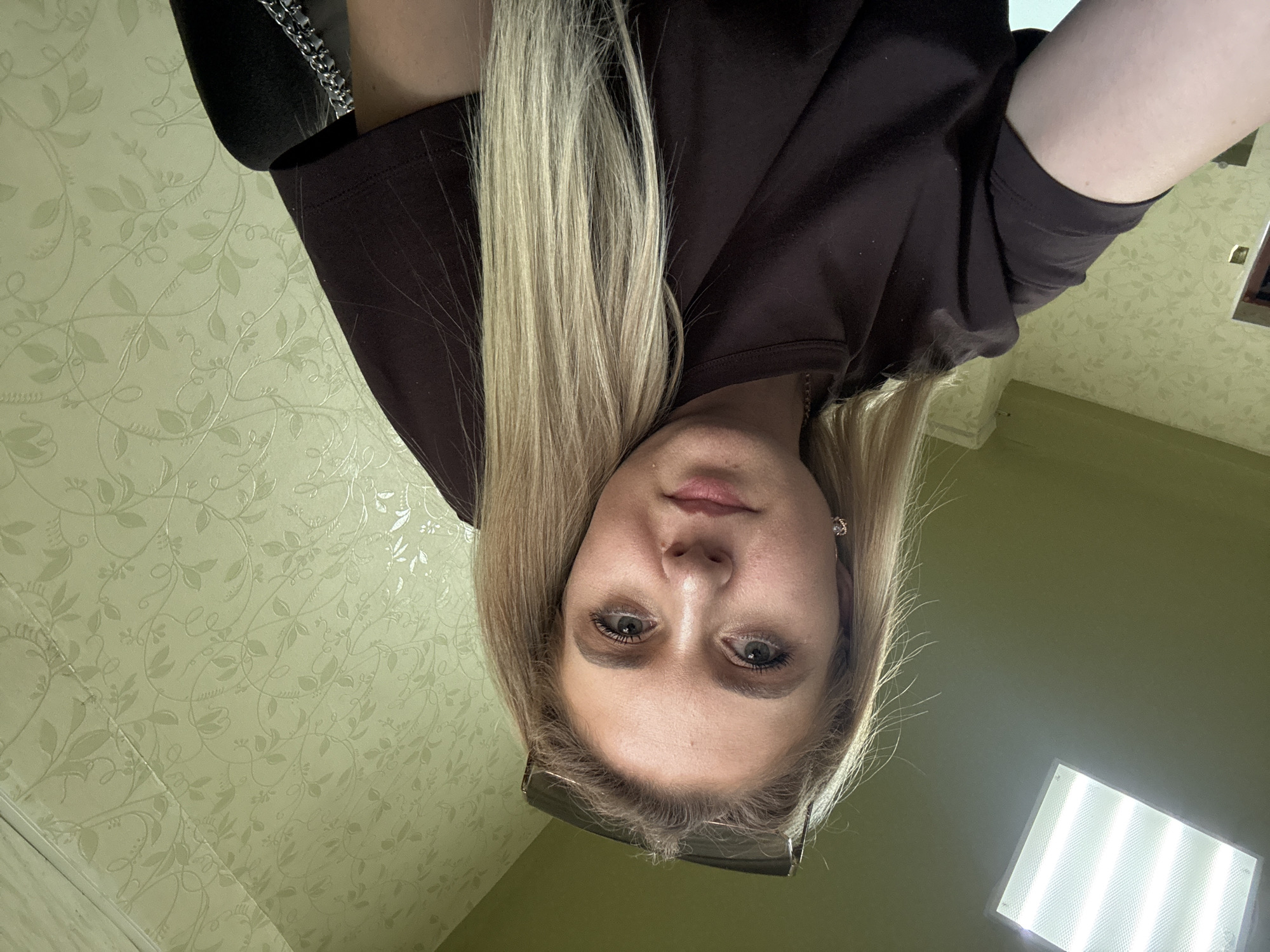 A woman with blonde hair and sunglasses perched on her head looks directly at the camera. She is wearing a dark brown t-shirt. The background consists of a pale green wallpaper with a floral pattern. A fluorescent light fixture is visible on the ceiling in the upper left corner of the image. The image is a selfie taken from a close-up perspective.