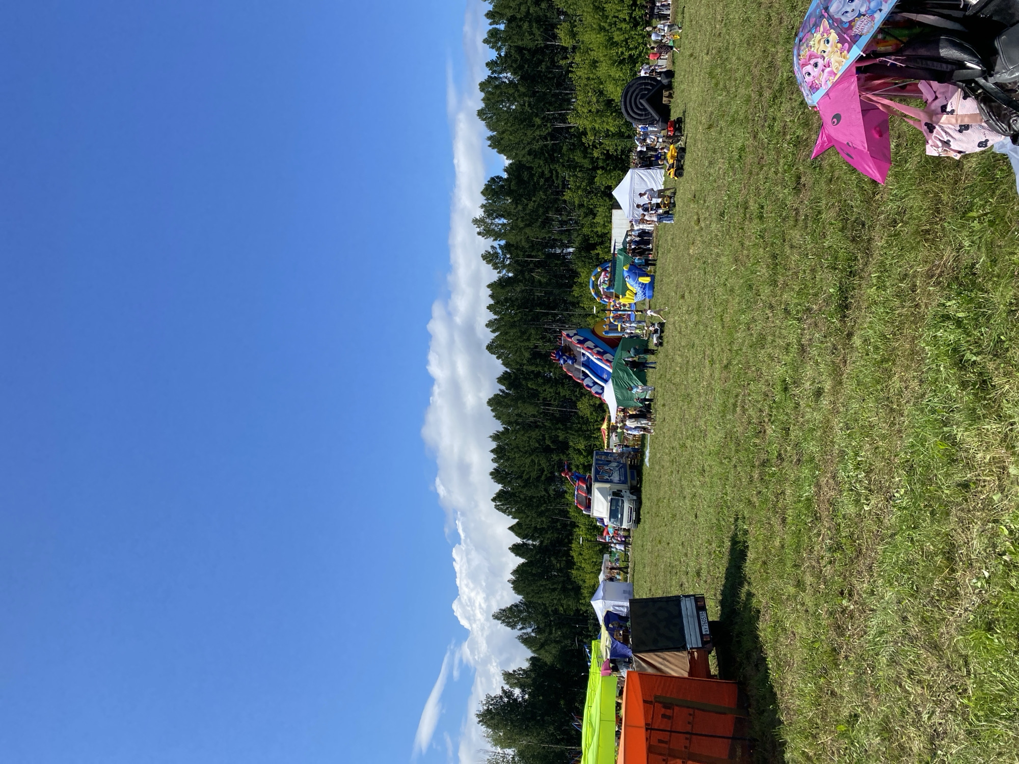 This image depicts a daytime outdoor event, likely a festival or fair, set in a grassy field with a dense line of trees in the background. The sky is a clear blue with scattered white clouds. In the foreground, to the right, are two open umbrellas, one pink and one decorated with cartoon characters, along with some bags. The middle ground of the scene is populated with various attractions. To the left, there are some large tent-like structures, one orange and one green, and a truck with a license plate visible. Further into the field, there are inflatable attractions, including a large blue and red slide and a colorful inflatable ride. Several white tents are also set up, and groups of people are seen milling about, some appearing to be children riding on small vehicles like ATVs. The overall atmosphere suggests a lively community event. The license plate on the truck appears to be from Russia. The presence of the license plate and the natural scenery are consistent with a location in Elovo, Russia.