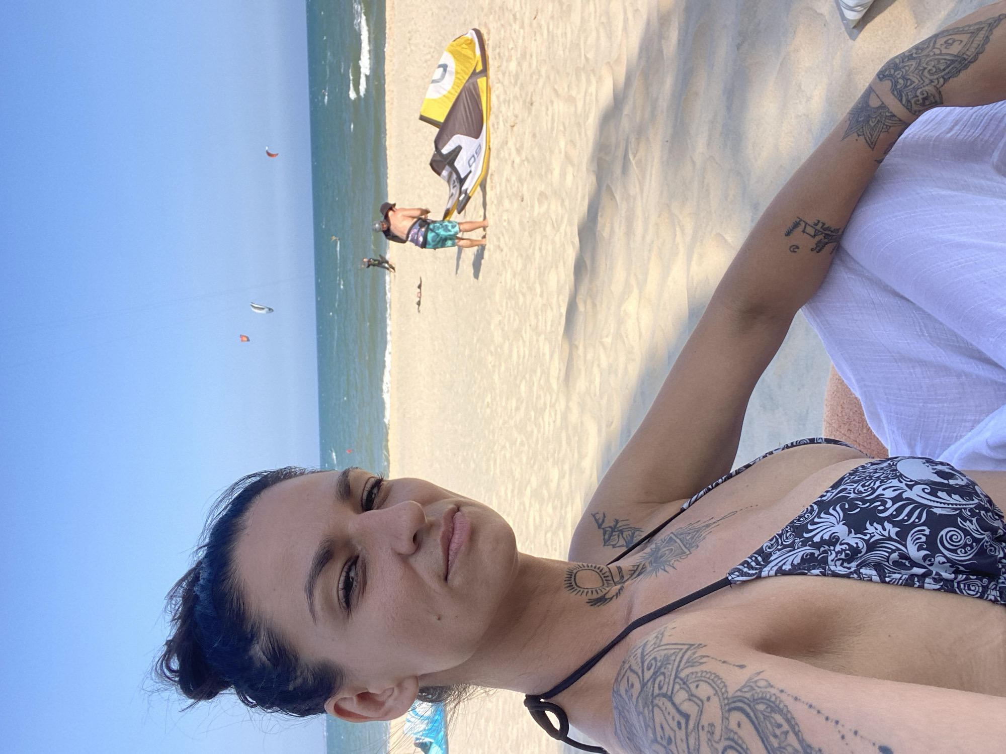 The image captures a sunny beach scene in Phu Thuy Ward, Vietnam. In the foreground, a Caucasian female with dark hair tied in a messy bun looks directly at the camera with a neutral expression. She wears a black and white patterned bikini top featuring damask and skull motifs, and a white sarong. Extensive tattoos are visible on her left shoulder, upper arm, right forearm, and neck (including a sun-like design). The light tan sand of the beach is visible. In the midground, an adult male wearing a dark top, patterned swim shorts, and a hat attends to a partially deflated yellow, black, and white kitesurfing kite on the sand. In the background, the turquoise-blue ocean features moderate waves and a red buoy. Several colorful kites (red, white, orange) are airborne, indicating kitesurfing activity further offshore under a clear, bright blue sky.