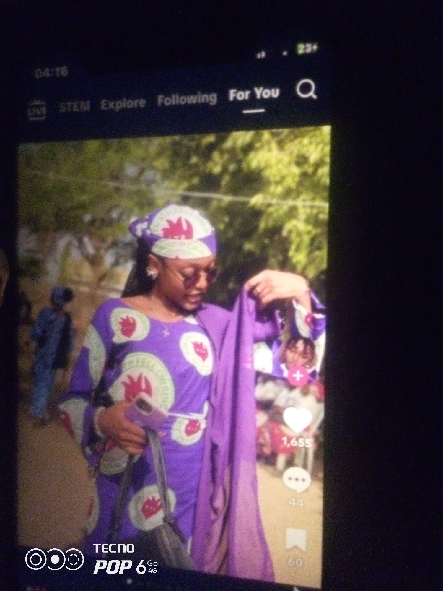 The image shows a young Black woman in the foreground, wearing a purple headscarf and matching long-sleeved dress adorned with a circular logo featuring a red stylized flame. She is also wearing round sunglasses and holding a purple cloth or garment. She appears to be holding a phone and a black handbag. In the background, blurred figures of other people are visible, as well as trees and greenery. The scene is captured on a screen, likely a mobile device, as indicated by the time stamp "04:16" and icons that resemble social media interface elements such as "LIVE," "STEM," "Explore," "Following," "For You," and a search icon. The bottom of the screen displays "TECNO POP 6 4G" branding. There is no indication of the location being in Howayi, Nigeria.