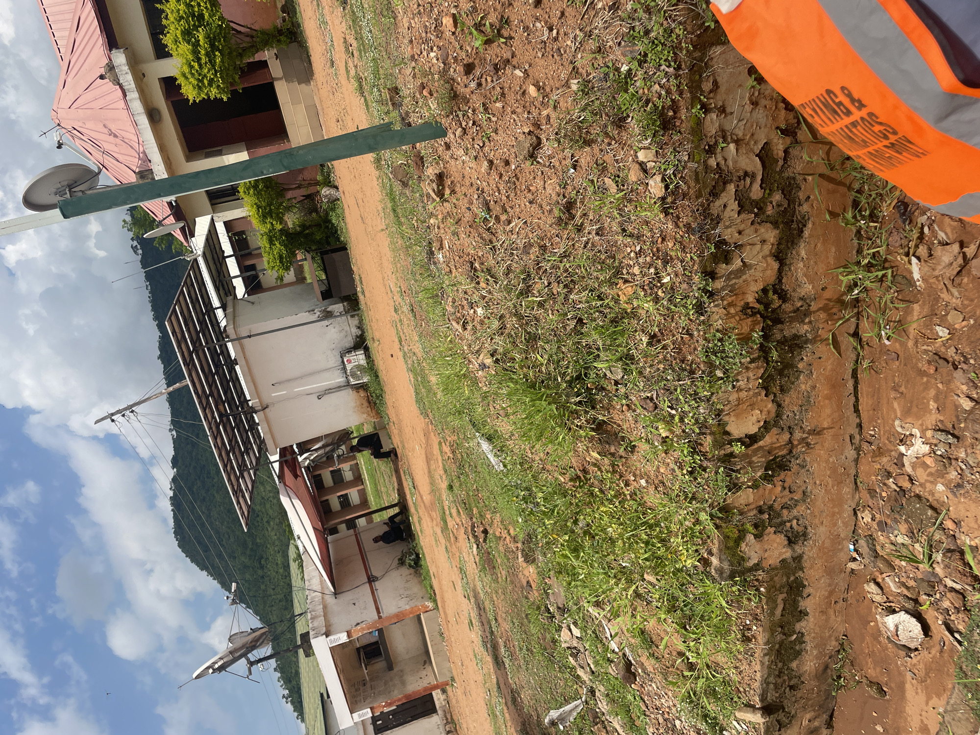 The image depicts an outdoor scene with a building and its surroundings. A portion of an orange safety vest with black text is visible in the top right corner. The text on the vest appears to read "SURVEYING & GEOMATICS". In the background, there is a building with a red-tiled roof and satellite dishes mounted on it. The building has a light-colored facade and appears to be situated on a slightly sloped terrain. Green trees are visible near the building.
The ground is covered with a mix of dirt, rocks, and sparse vegetation, including grass and some small wildflowers. A trench or ditch with some water is present in the foreground, along with mossy patches on its edges. The sky is cloudy, suggesting overcast weather. The scene is likely in Lokoja, Nigeria, as indicated by the provided context. The overall impression is of an undeveloped or rural area adjacent to a building. No specific activity or event is clearly depicted, apart from the suggestion of surveying work due to the vest. The time of day is not explicitly indicated, but the lighting suggests it is daytime.