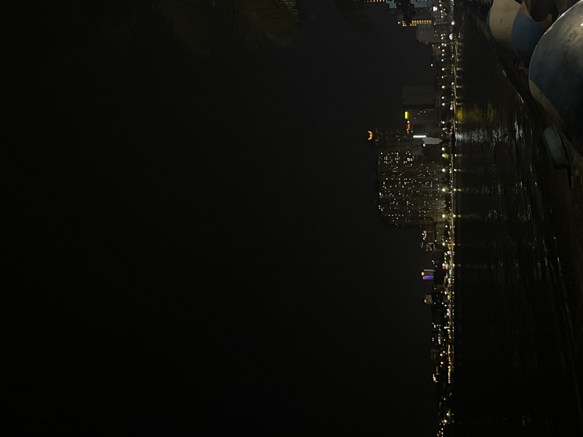 The image depicts a nighttime view of a coastal city, likely Bac Nha Trang Ward, Vietnam. In the foreground, the dark water of the sea reflects the city lights. On the right side, partially visible, are boat-like objects covered in white and blue material, suggesting they are moored or undergoing maintenance. The middle ground showcases a brightly lit cityscape with numerous buildings and skyscrapers. The prominent building in the center features a distinctive crescent moon-shaped illumination on its roof. The sky above is dark, with only the illuminated city providing light. The overall scene captures the vibrant night life of a coastal urban area.