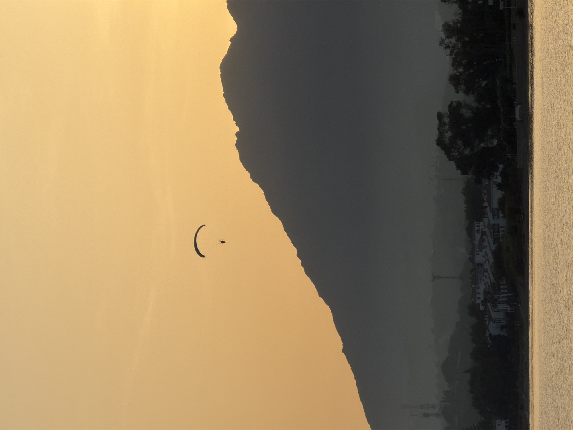 A paraglider is silhouetted against an orange sky over a dark, mountainous landscape in Estepona, Spain. Below the mountain, a village with white buildings is visible, nestled among dark trees. In the foreground, a body of water reflects the warm light of the sky. The scene appears to be a sunset or sunrise.