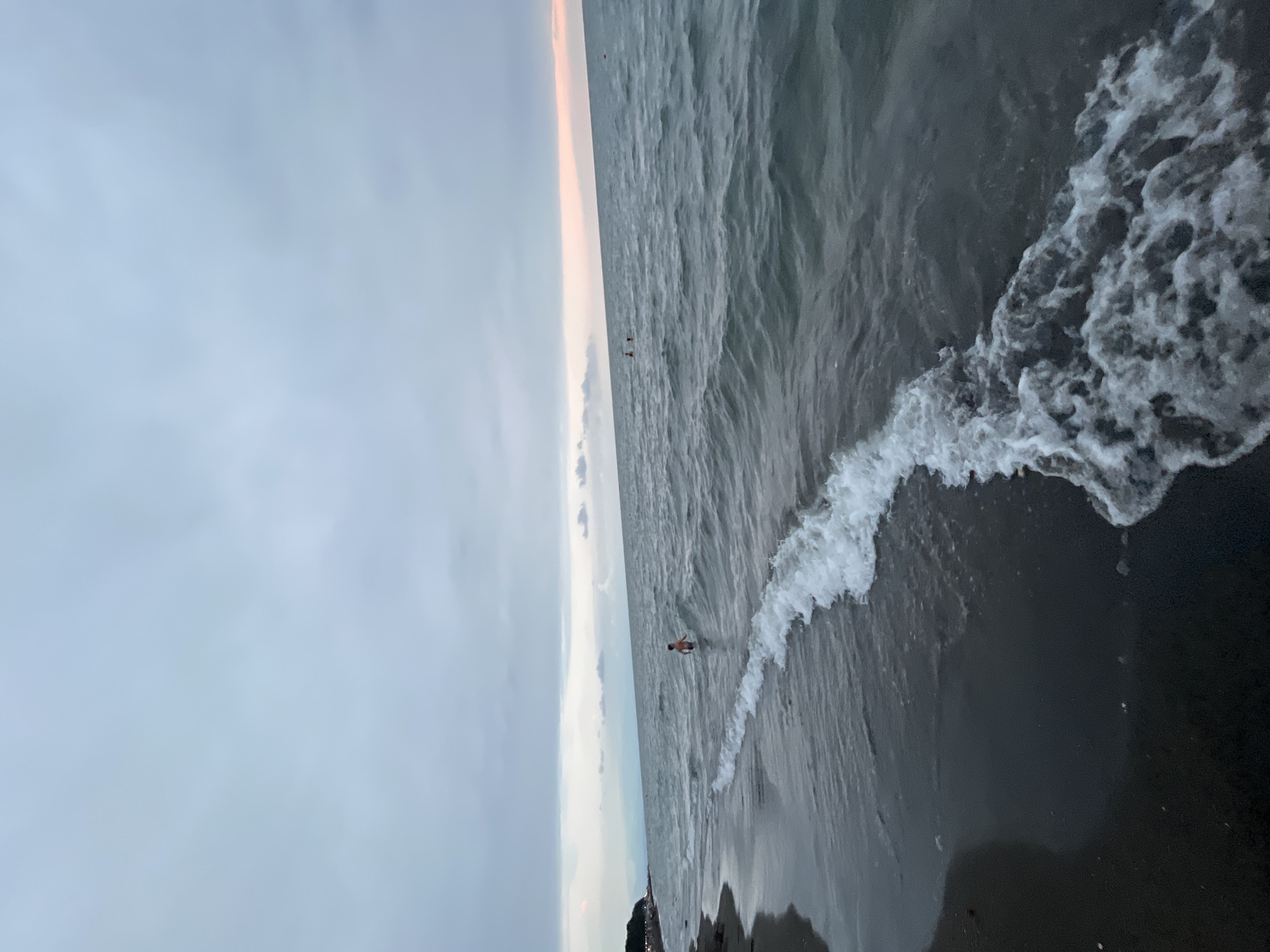 The image depicts a beach scene in Ureki, Georgia, characterized by a dark sandy shore meeting a gray, choppy sea under an overcast sky. In the mid-ground, a child stands in the shallow water, facing away from the camera. Further out in the sea, two small figures can be seen, possibly swimmers, and a red marker buoy is visible on the horizon. The water exhibits a dark, greenish-gray hue, with white foam from breaking waves washing onto the shore. The distant coastline features a strip of greenery and structures, suggesting a populated area. The overall mood is subdued, likely due to the cloudy weather.