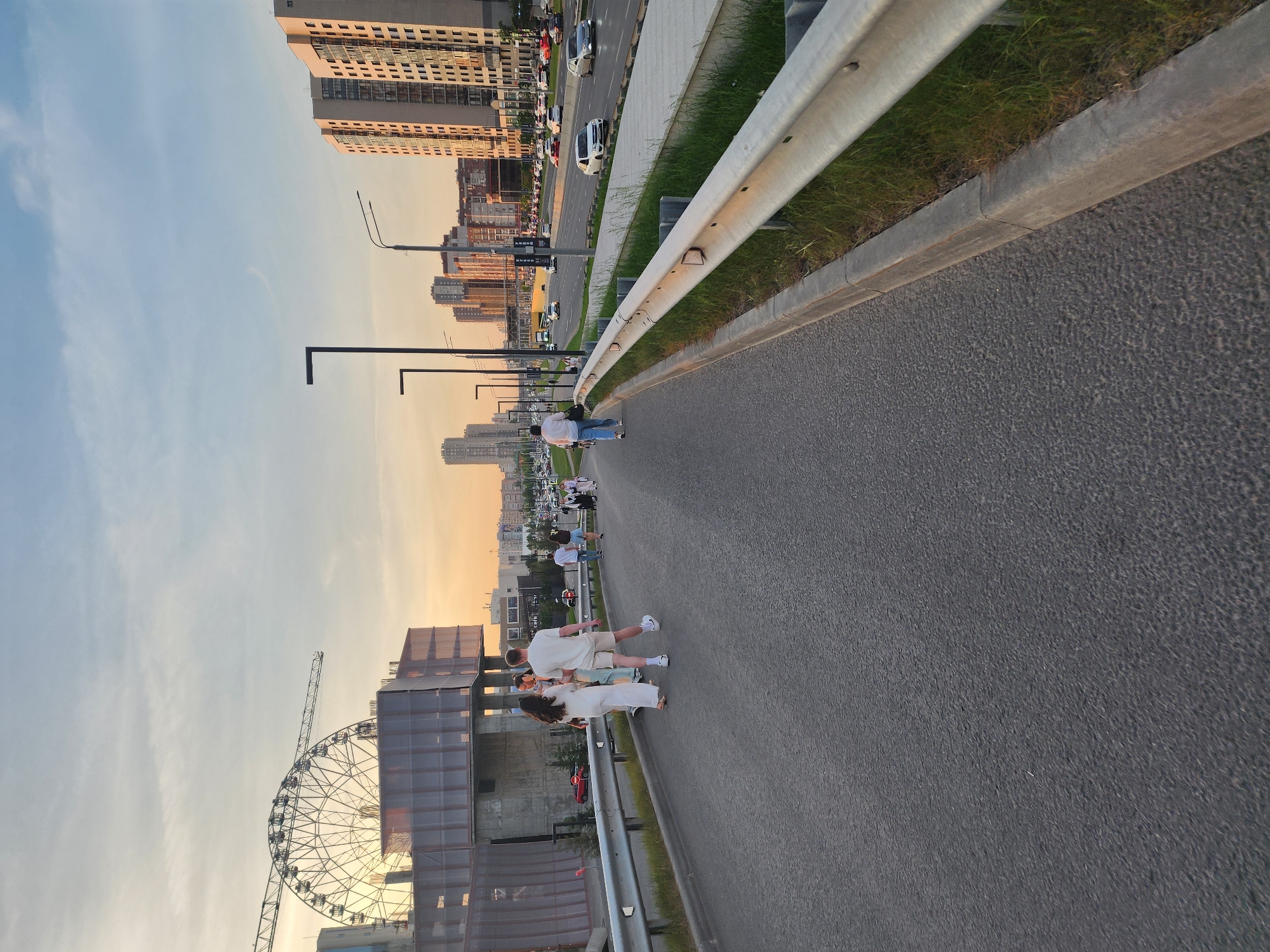 The image shows a couple walking along a pedestrian walkway next to a road in Kazan, Russia, with the Ferris wheel visible. The road features a metal guardrail, grass, and trees. There are other people walking on the sidewalk in the distance. The sky is mostly blue with some orange tones near the horizon. Buildings are in the background on the left and right. Cars and pedestrians can be seen on the road in the distance. A couple is holding hands.