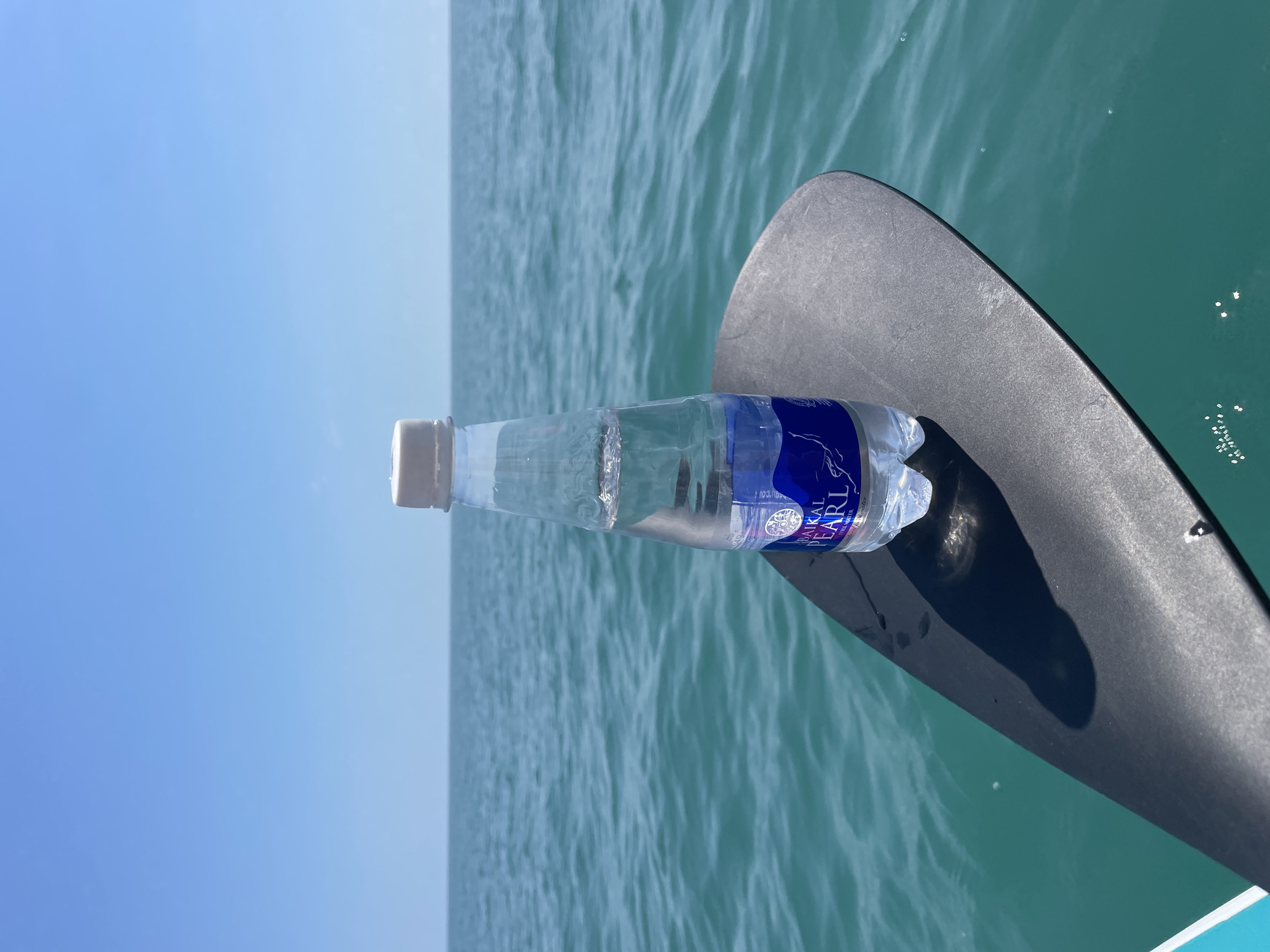An over-the-shoulder view captures a clear plastic bottle of water with a white cap, resting on a black paddle. The bottle, branded "Baikal Pearl Still Water" with a blue and white label, is half full. The paddle is positioned in the foreground, extending from the bottom left corner towards the right. Beyond the paddle, a vast expanse of calm, turquoise water stretches to the horizon, meeting a clear blue sky. The scene suggests an outdoor activity, possibly on a body of water. The location is identified as Sochi, Russia.
