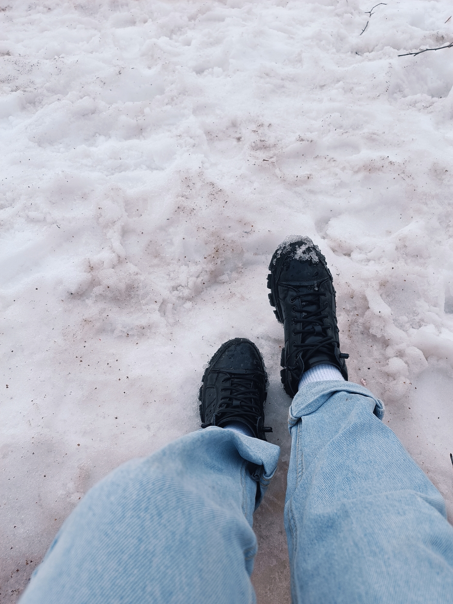 A first-person perspective photograph shows a pair of legs in light blue jeans and black boots standing on dirty snow. The snow is melting and contains dirt and debris. The location is purported to be Izhevsk, Russia.