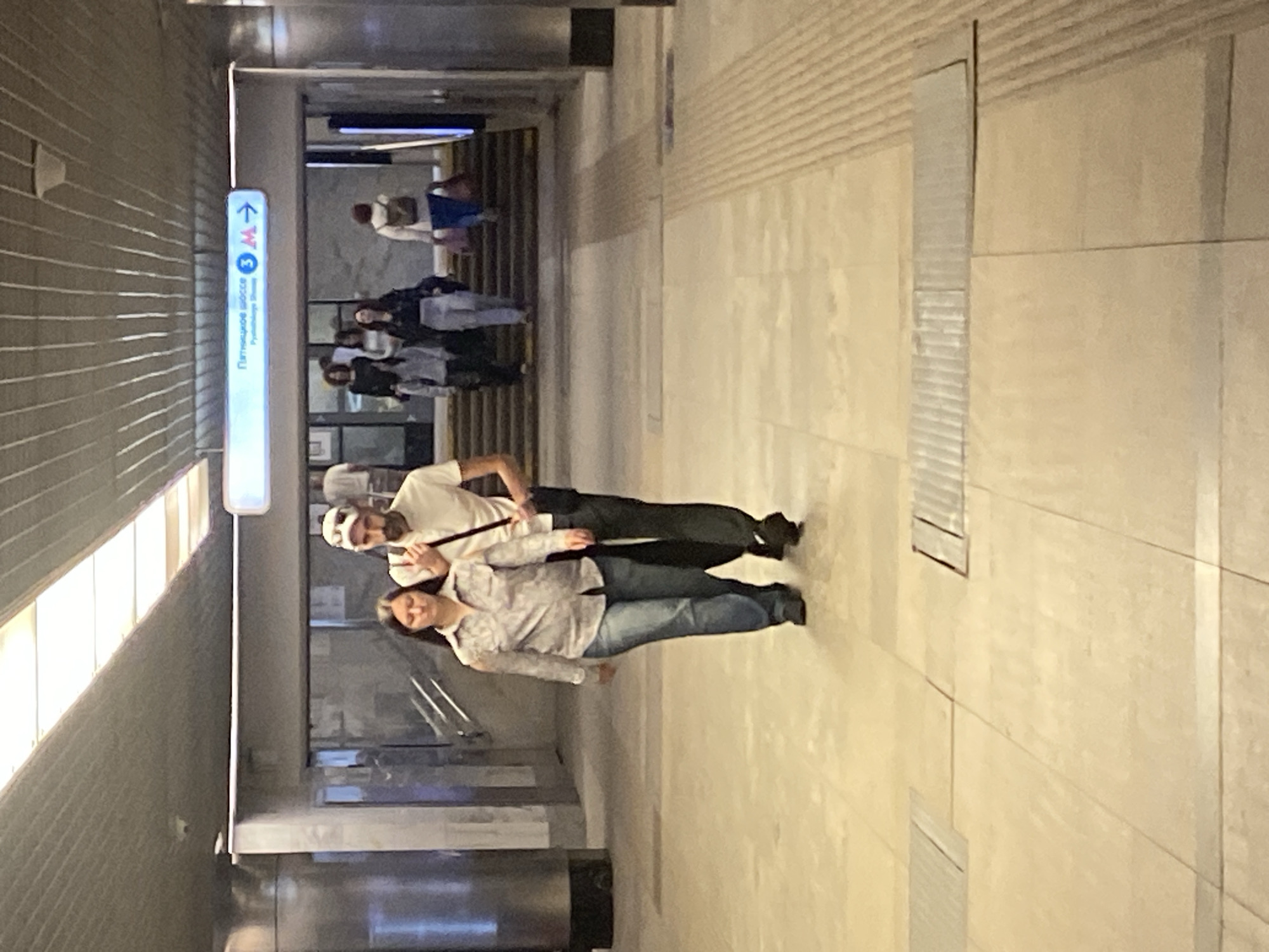 The image depicts an indoor scene, likely a subway station or underpass in Khimki, Russia, given the signage. In the foreground, a couple is walking towards the viewer. The woman on the left is wearing a patterned long-sleeved shirt and blue jeans. The man on the right is wearing a white short-sleeved shirt, dark pants, and a white baseball cap. He has a dark bag slung over his shoulder. Behind them, other people are seen walking up a set of stairs towards a metro station entrance, indicated by directional signage with Russian text and the Moscow Metro logo. The environment features a polished, light-colored tiled floor with textured strips for tactile guidance. The ceiling is constructed with long, narrow metallic panels, and fluorescent lighting is visible. The overall impression is one of a functional, modern public transportation hub.