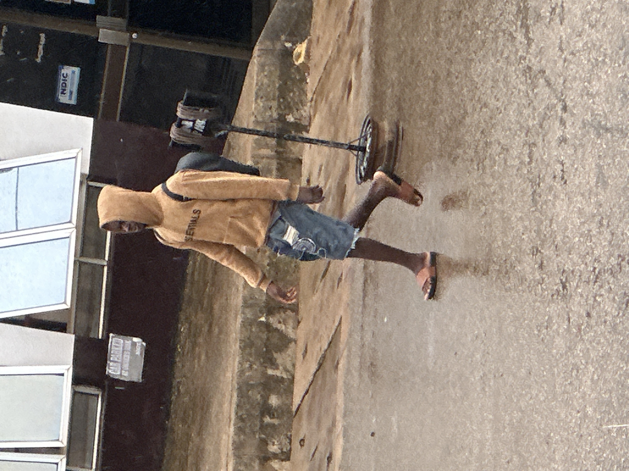 A person wearing a light orange hooded sweatshirt with "ESSENTIALS" printed on the front and denim shorts walks on a wet asphalt surface in Bukuru, Nigeria. The person is carrying a backpack and wearing sandals. In the background, there is a building with windows and a sign that reads "NDIC". A metal pole with a circular base stands on the sidewalk. The ground is wet, indicating it might have rained recently.