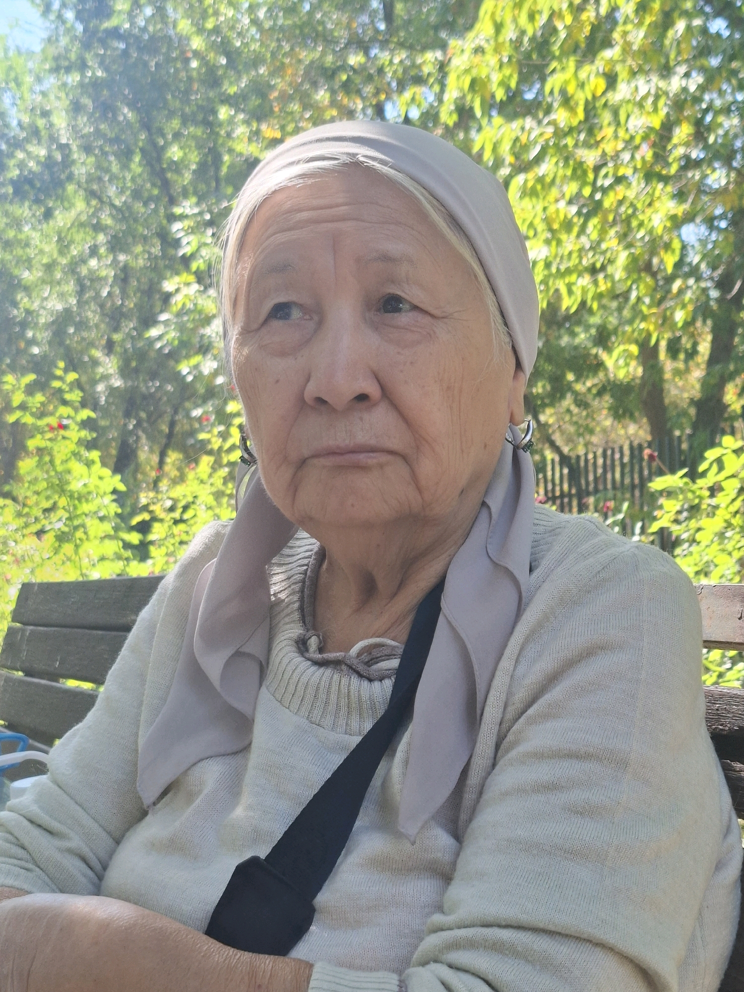 A medium shot captures an elderly East Asian woman from the chest up, seated on a wooden park bench. She is wearing a light-colored, long-sleeved sweater and a pale headscarf that drapes over her shoulders. A black strap crosses her chest from left to right, suggesting a bag is present. Her expression is neutral, with her gaze directed slightly off-camera to the left. The background consists of blurred greenery from trees and foliage, with a hint of a wooden fence. The lighting suggests it is daytime, likely outdoors in a park. The scene does not provide specific indicators to pinpoint a location within Almaty, Kazakhstan, but it depicts a typical park setting that could be found in many urban areas.