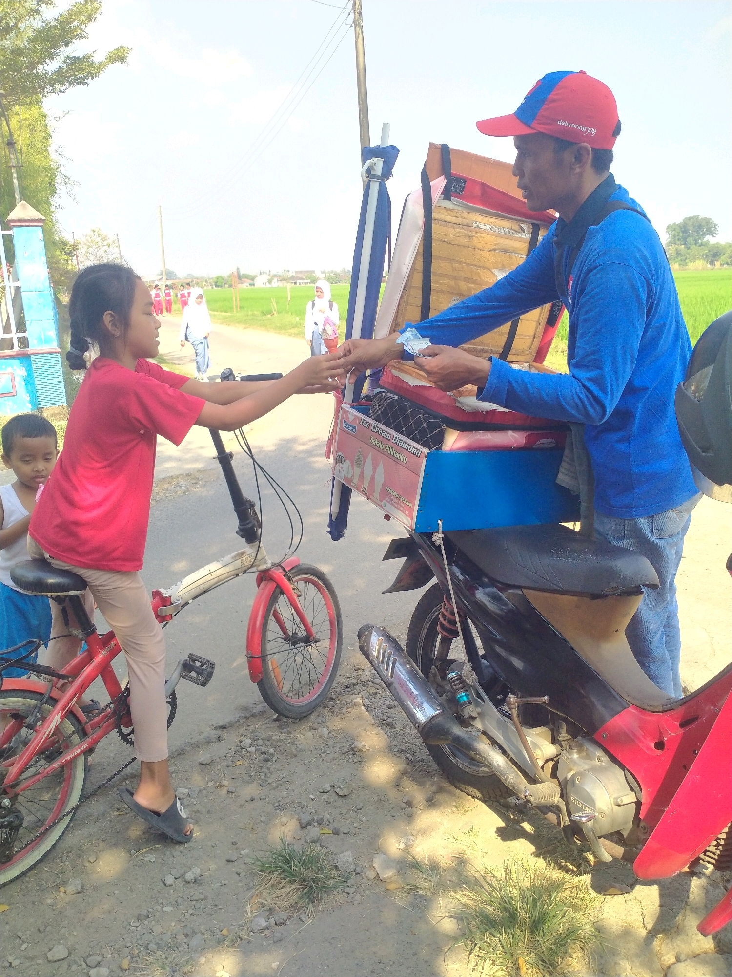 In Begajah, Indonesia, a young girl wearing a red shirt and beige pants sits on a red bicycle, reaching out to a man. The man, wearing a blue long-sleeved shirt, a red and blue baseball cap, and blue jeans, stands beside a motorcycle with an ice cream cart. He is handing a small item, likely money or an ice cream, to the girl. Behind them, other children and adults are visible in the background, some walking on a dirt road. The environment is rural, with fields of green crops and trees surrounding the scene under a bright sky. The ice cream cart is decorated with images of ice cream and text that reads "Ice Cream Diamond".
