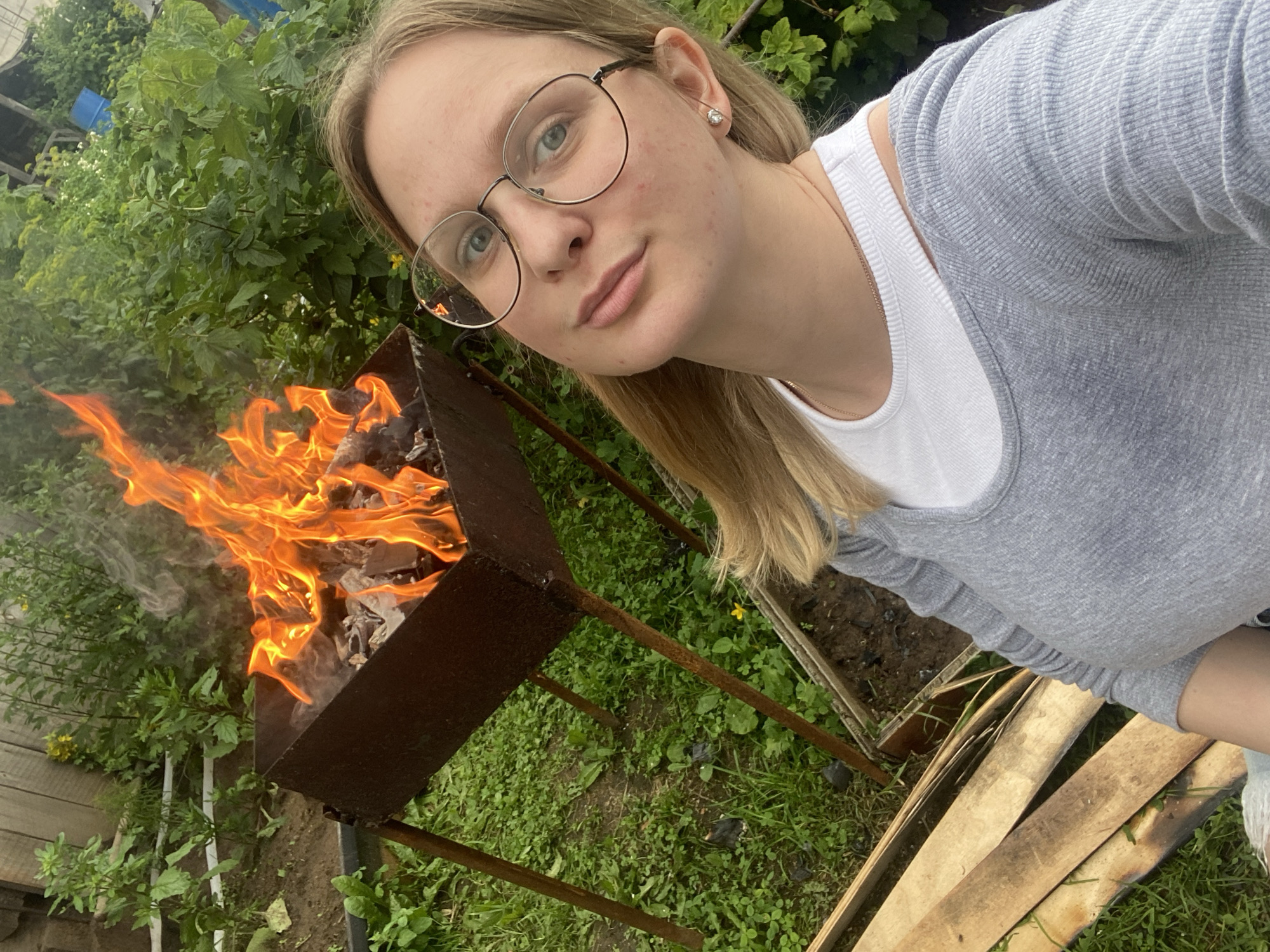 In Kauchuk Stantsiya, Russia, a young woman with blonde hair and glasses is taking a selfie outdoors. She is wearing a grey long-sleeved shirt over a white t-shirt and a necklace. Behind her, a barbecue grill is on fire, with flames and smoke rising from it. The grill is situated on a grassy area with some wooden planks in the foreground and lush green foliage in the background.