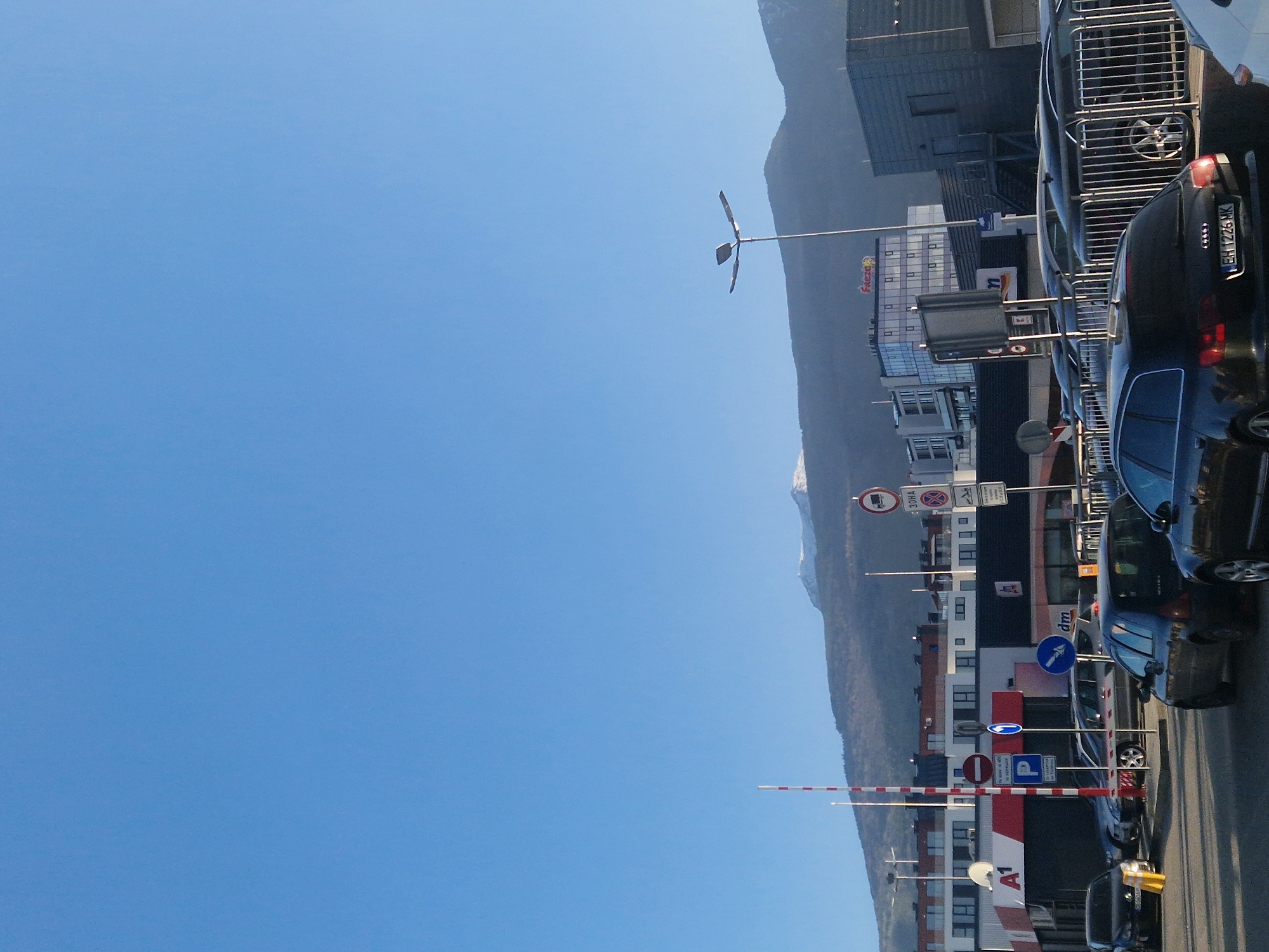 The image depicts an urban street scene set against a backdrop of a large mountain range under a clear blue sky. The photo is taken on a bright, sunny day, likely midday, with excellent visibility.
In the background, a substantial mountain range stretches horizontally across the frame, with some peaks exhibiting patches of snow, suggesting a cooler climate or higher elevation. The sky above is a uniform, clear blue, devoid of clouds.
The foreground and mid-ground are dominated by a street lined with buildings and parked vehicles. On the left side, several cars are parked, mostly dark-colored. The rear of a black car is clearly visible, displaying a license plate that reads "EH 1226 XK". Various other cars, including a white one, are parked further along the street.
Several multi-story buildings, featuring modern architectural styles with numerous windows, line the street. These buildings host commercial establishments. Visible logos and text on the buildings include "Frezza", "dm" (identifying a drugstore), and "A1" (a telecommunications company).
The street is densely populated with street furniture and signage. A distinct street light with a multi-pronged fixture stands tall. Numerous traffic signs are present, including a red circle with a white bar (no entry), a blue circle with a white "P" (parking), directional arrows (blue circles with white arrows), and other regulatory signs like "30HA". Red and white striped barrier arms are also seen, possibly indicating an entrance or restricted area. Some signs contain text in Cyrillic script, like "СЪОБРАЗНО М", suggesting an Eastern European location.
No people are visible, and no specific activity or event is taking place; it's a static capture of a bustling street environment in a mountainous region.